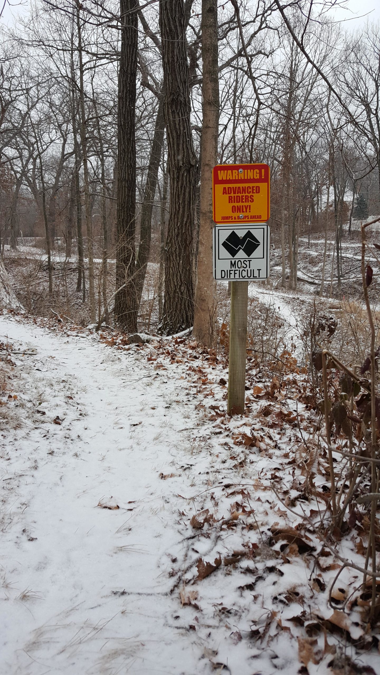 A snowy path through a wooded area with two warning signs. The first sign reads "WARNING! ADVANCED RIDERS ONLY! JUMPS & DIPS AHEAD," while the second sign indicates the trail is "MOST DIFFICULT." The ground is covered with a light layer of snow and fallen leaves. Memorial Park Trails mountain bike trail.