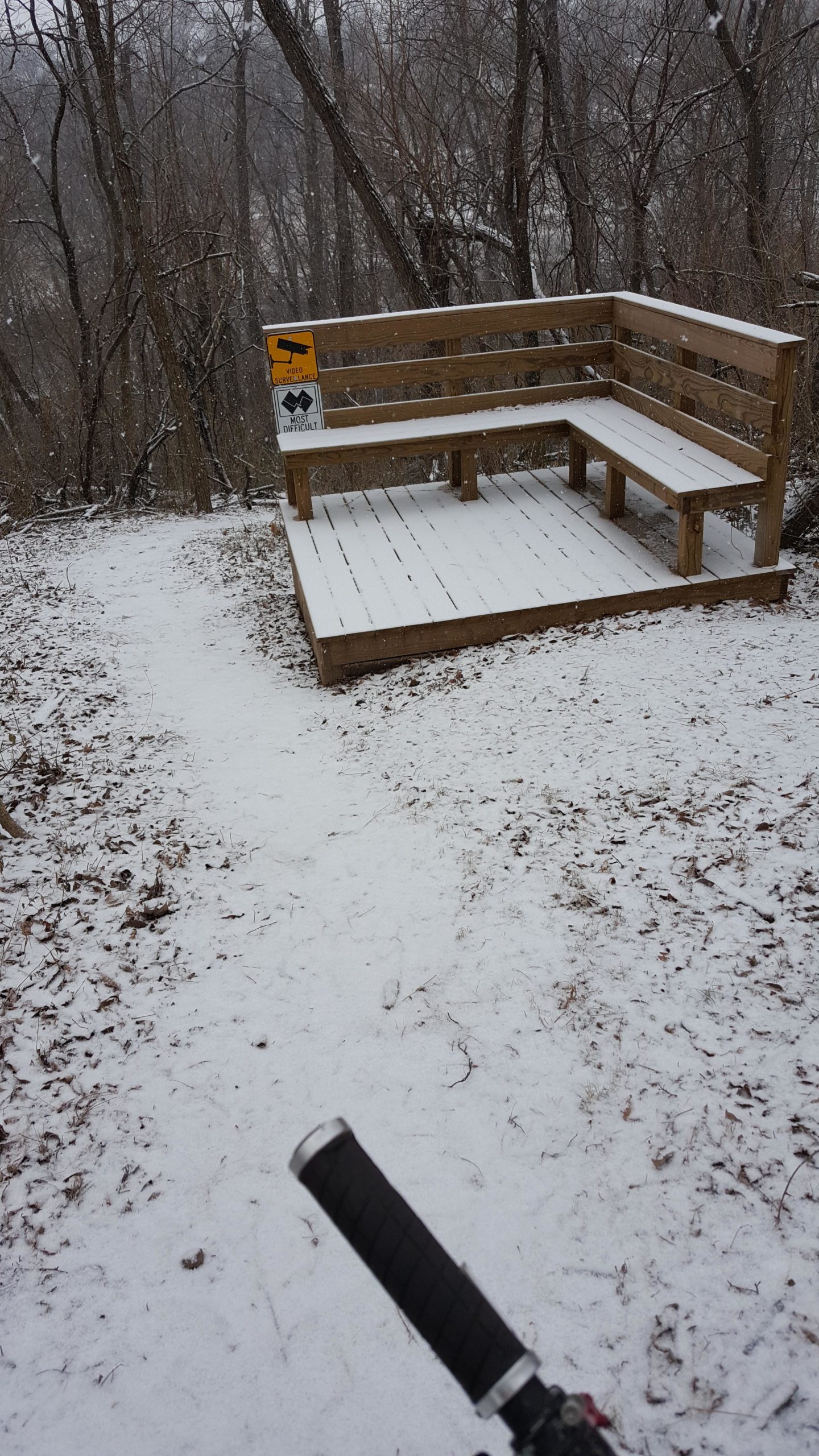 A wooden observation deck covered in snow, located along a winding trail surrounded by bare trees. A sign indicates a difficult trail ahead. A portion of a bicycle grip is visible in the foreground, suggesting an outdoor adventure context. Memorial Park Trails mountain bike trail.