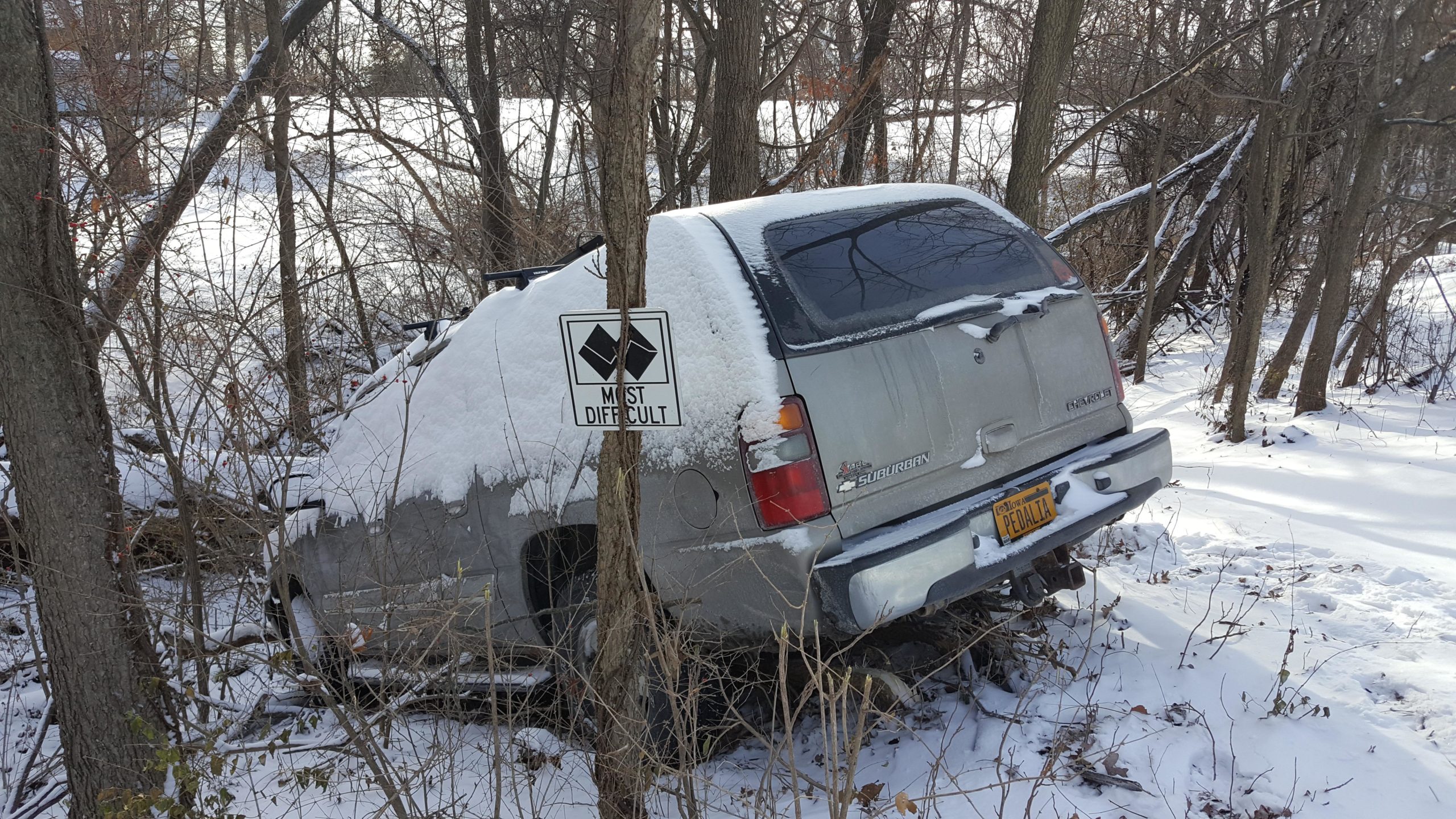An abandoned silver SUV partially covered in snow is situated among trees in a snowy landscape. A warning sign labeled "MOST DIFFICULT" stands next to the vehicle, which is partially obscured by branches and underbrush. Memorial Park Trails mountain bike trail.