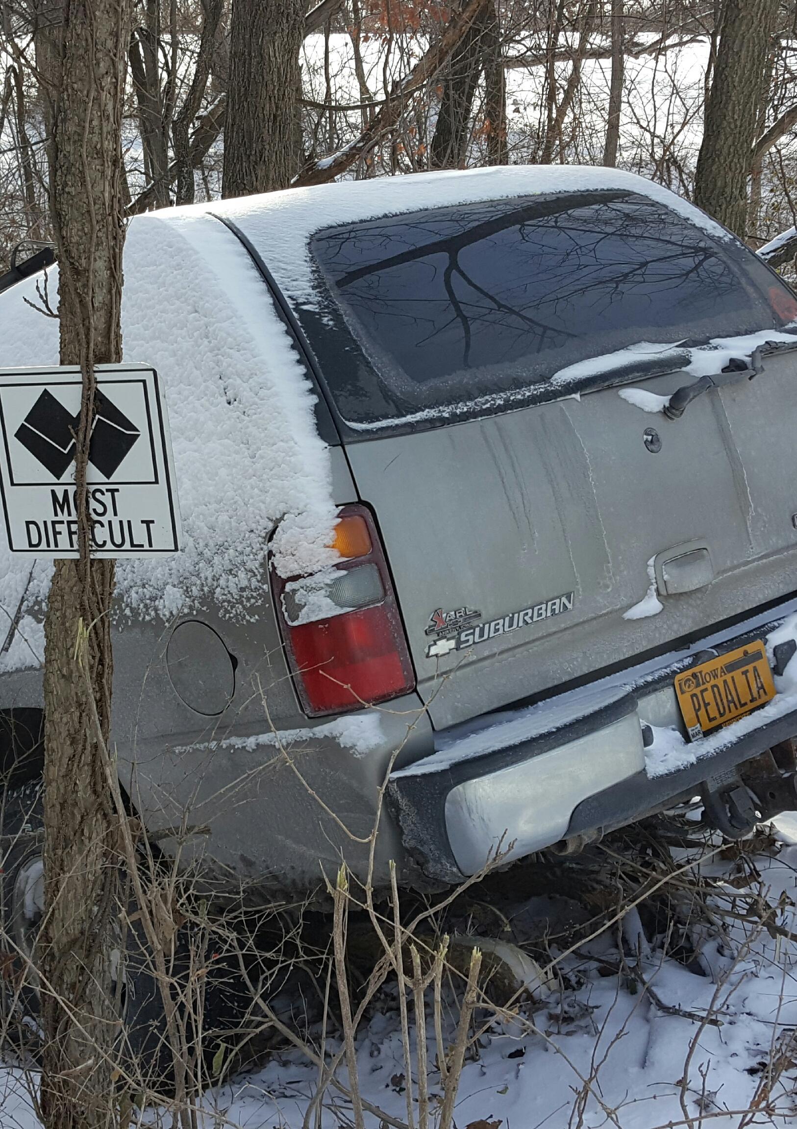 A silver Chevrolet Suburban covered in snow and ice is partially hidden among trees and brush. A sign next to the vehicle indicates "Most Difficult." The rear of the Suburban is visible, showcasing its brand logo and a license plate that reads "PEDALIA." Memorial Park Trails mountain bike trail.