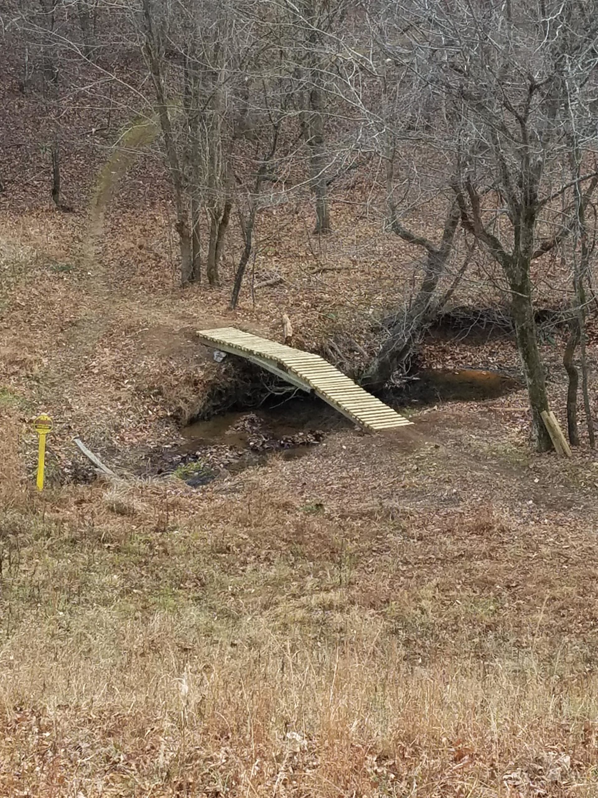 A wooden footbridge spans a small creek in a wooded area. The surrounding landscape is covered in fallen leaves and dry grass, with bare trees visible in the background. A yellow trail marker is positioned nearby, indicating a hiking path. Dark Hollow mountain bike trail.