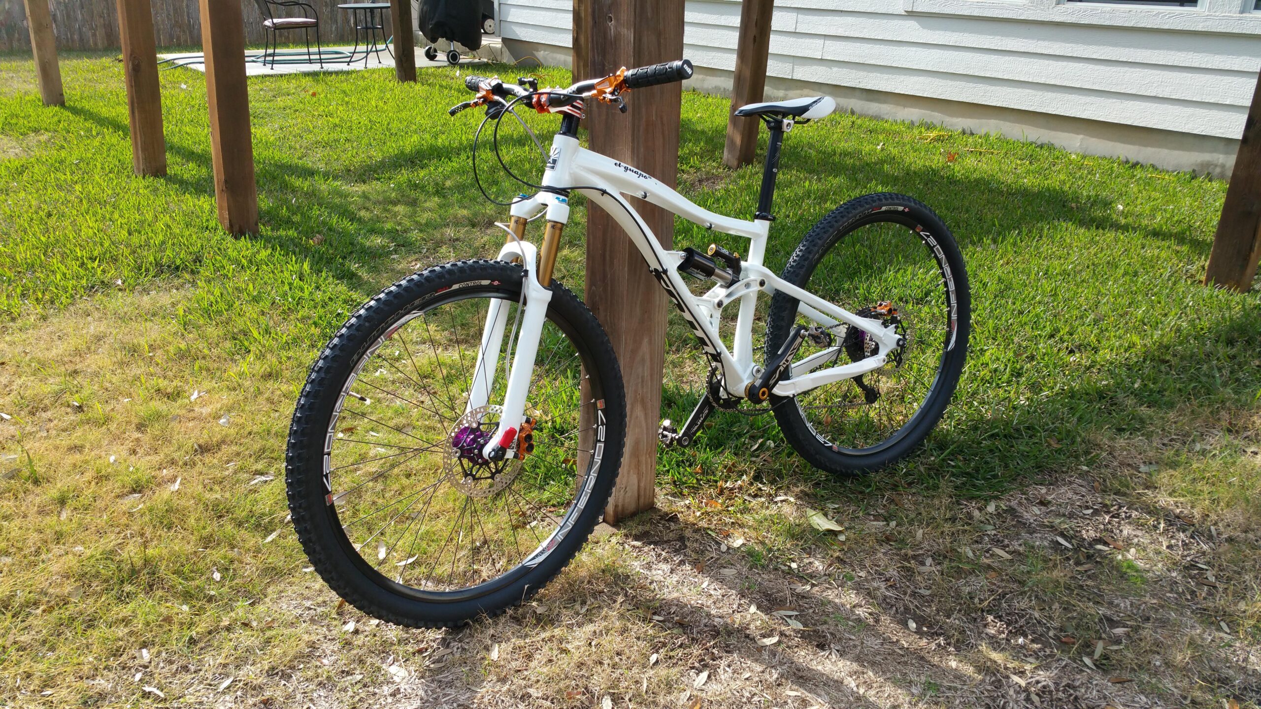 Titus El Guapo: A white mountain bike with black tires and purple and orange components is leaning against a wooden post in a grassy area. In the background, there is a deck with a table and chairs, along with a white house.