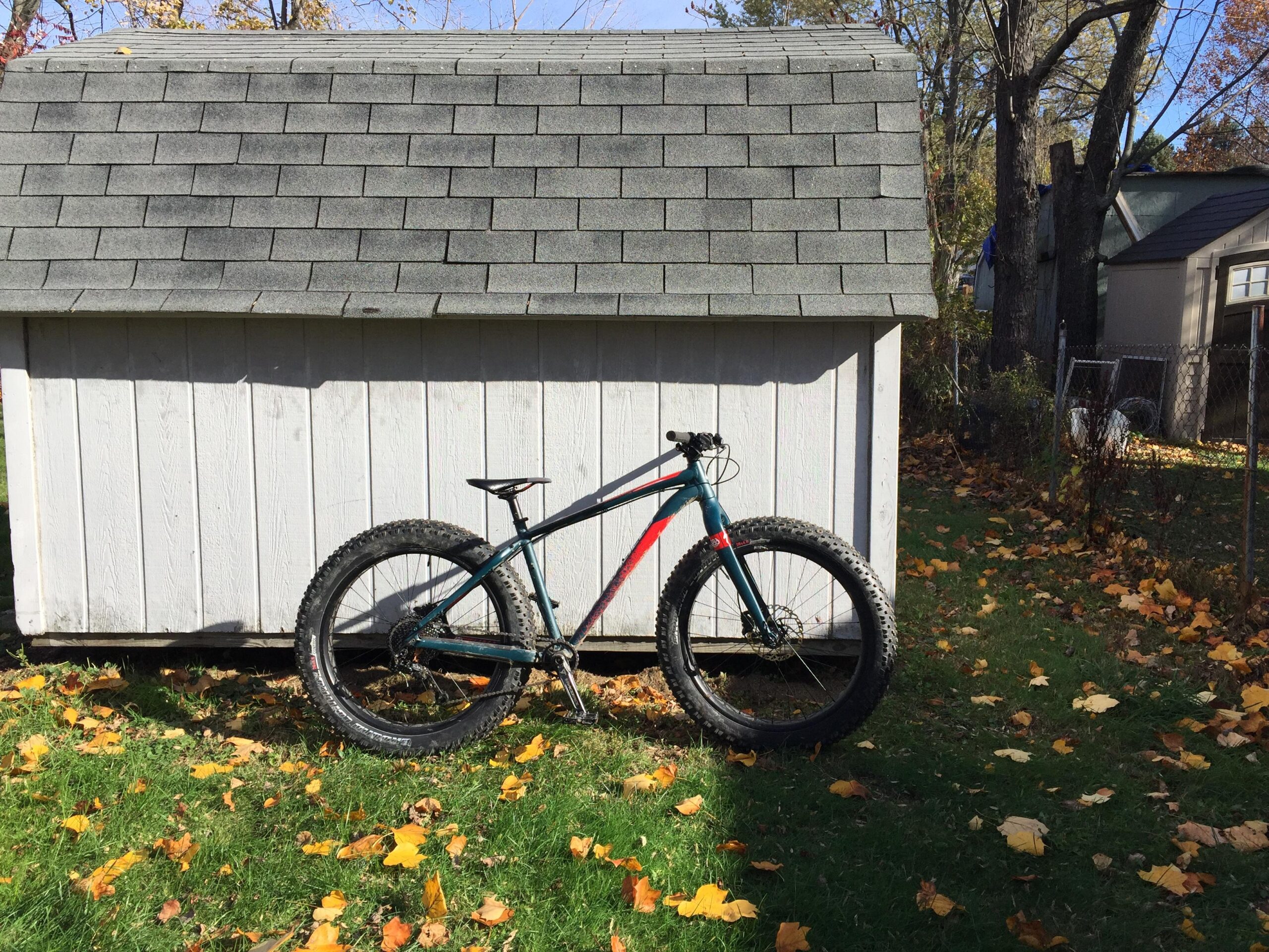 Specialized Fatboy Pro: A fat bike with large tires is parked beside a white wooden shed, surrounded by green grass and fallen yellow and brown leaves on the ground. The background features trees and additional structures, with a clear blue sky above.