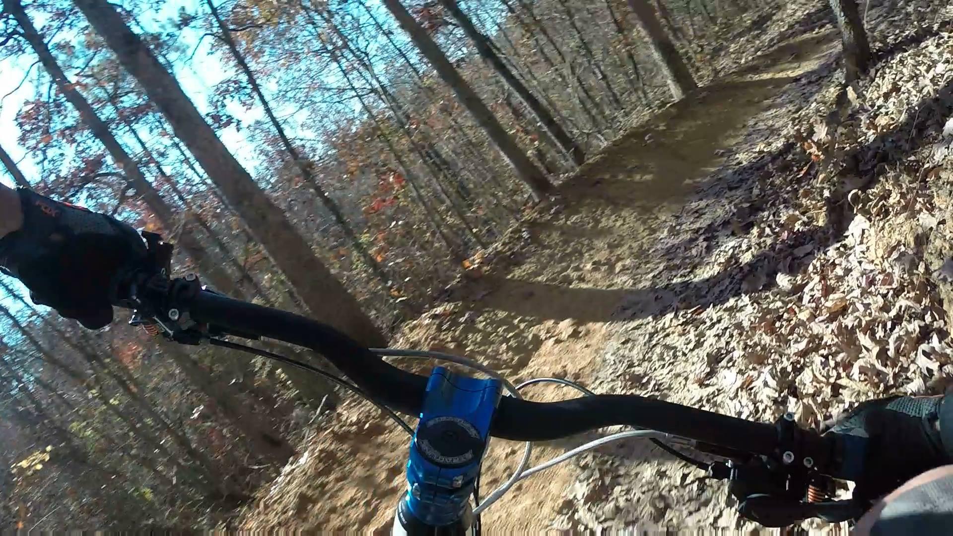 A close-up view of a mountain bike handlebar with a gloved hand gripping it, navigating a dirt trail in a wooded area. The scene captures autumn foliage with brown and orange leaves on the ground and trees lining the trail under a clear blue sky. Taylor Randahl Memorial Mountain Bike Trails At Olde Rope Mill Park mountain bike trail.
