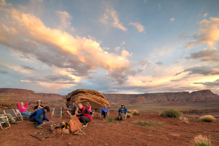 A group of people gathered around a campfire in a desert landscape, seated in reclining chairs. The scene is set against a backdrop of red rock formations and a colorful sunset sky with scattered clouds. Some individuals are smiling and engaging with one another, surrounded by the natural beauty of the desert.