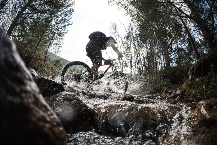 A mountain biker rides through a rocky stream, creating splashes of water as he navigates the terrain. The scene is set in a wooded area with trees in the background and a cloudy sky above, capturing the dynamic and adventurous spirit of outdoor biking.