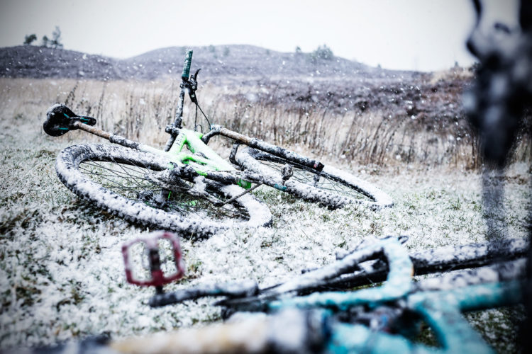 A mountain bike rests on the ground, covered in snow, with falling snowflakes in the foreground. The scene captures a winter landscape with muted colors and blurred hills in the background.