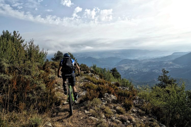 A cyclist rides along a rocky mountain trail surrounded by vegetation, with a panoramic view of valleys and hills in the background under a partly cloudy sky.