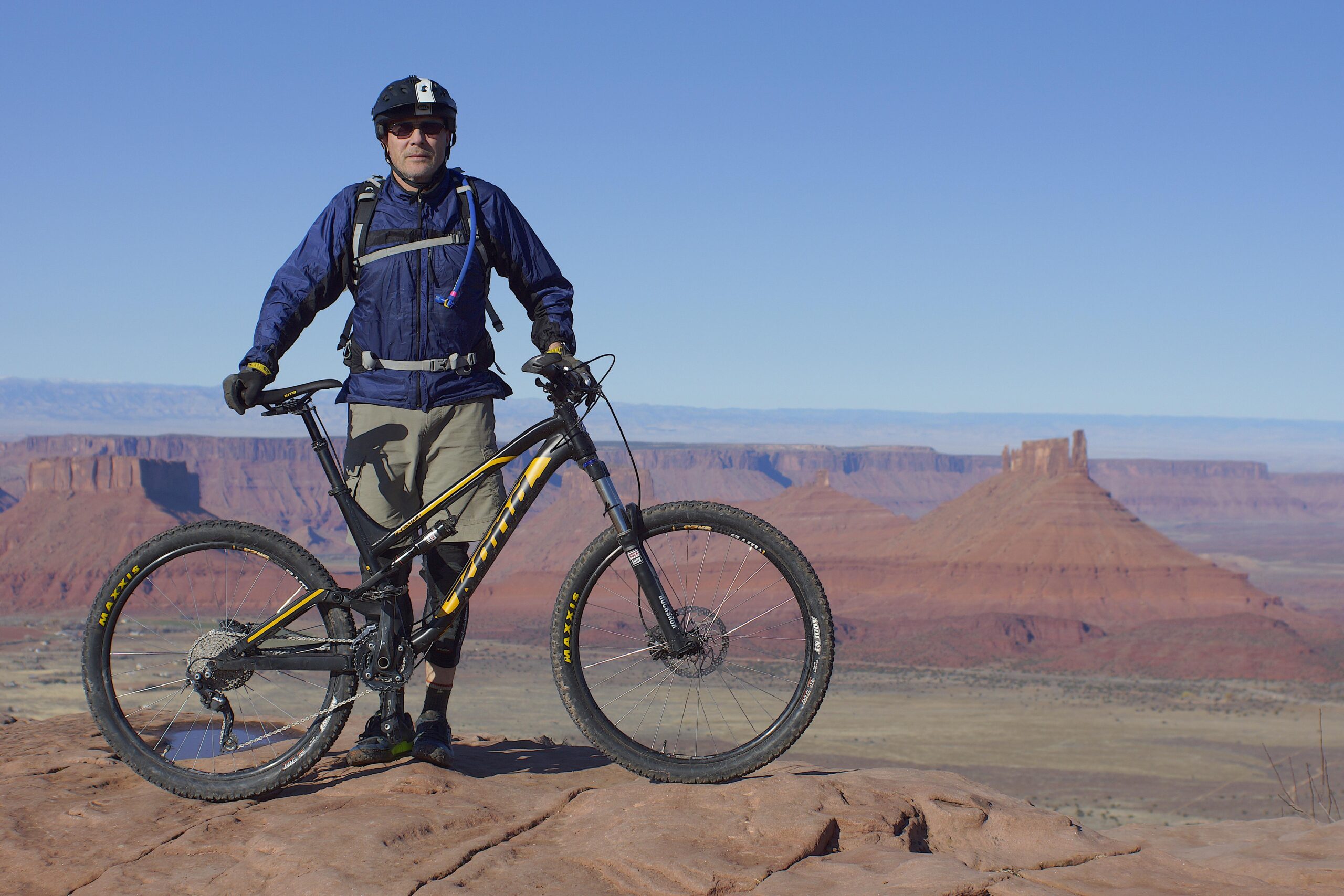 Kona Process 134: A mountain biker wearing a helmet and a blue jacket stands on a rocky ledge with a black and yellow mountain bike beside him. In the background, red rock formations and mountains stretch across a clear blue sky. The scene captures the essence of outdoor adventure in a rugged landscape.