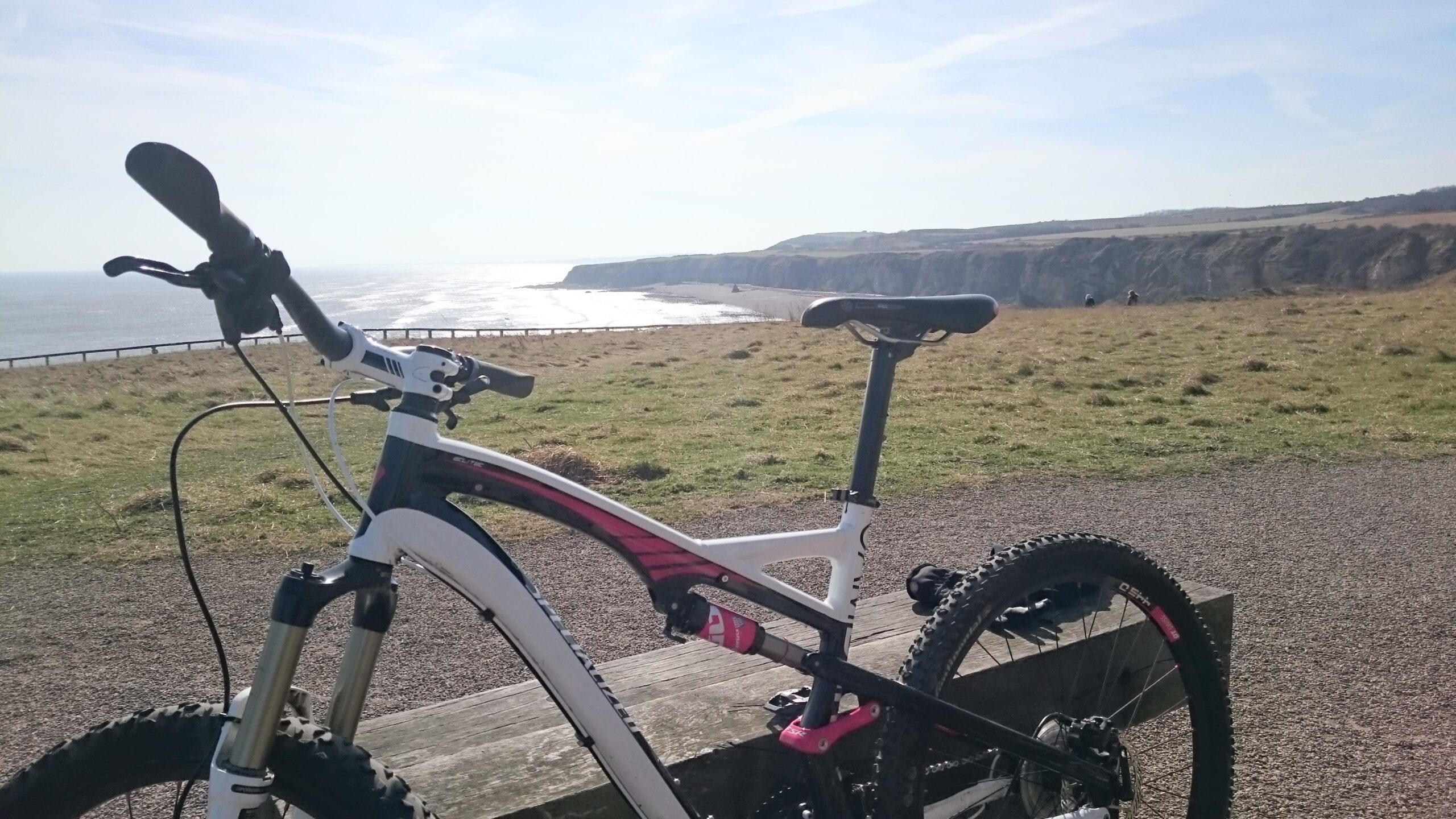 Specialized Camber: Mountain bike resting on a wooden bench with scenic coastal cliffs and the ocean in the background on a bright, sunny day.
