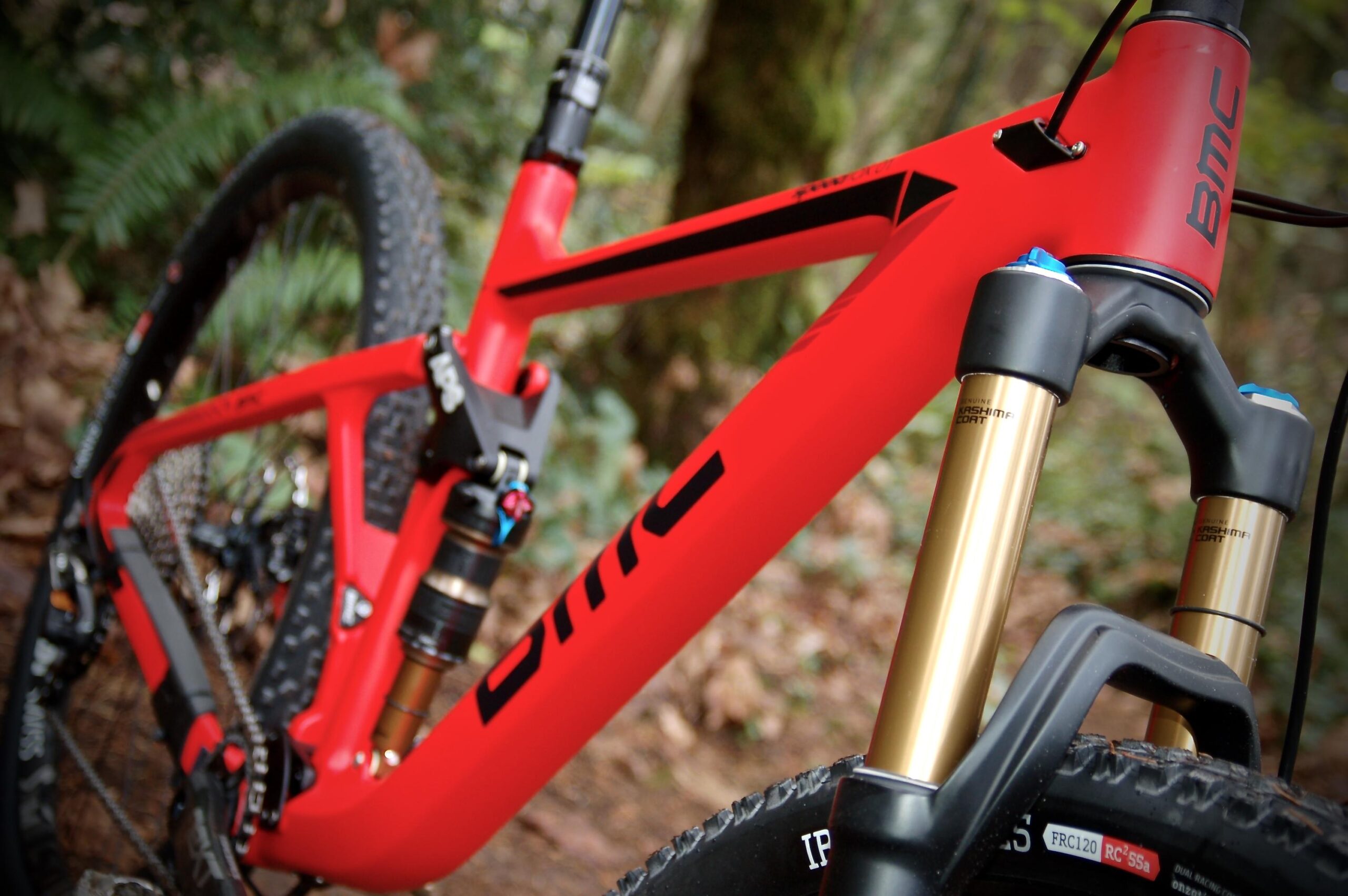 BMC Speedfox 01: A close-up image of a red mountain bike frame displaying the BMC logo, featuring a gold suspension fork and details of the drivetrain. The background includes blurred greenery and foliage, suggesting an outdoor, woodland setting.