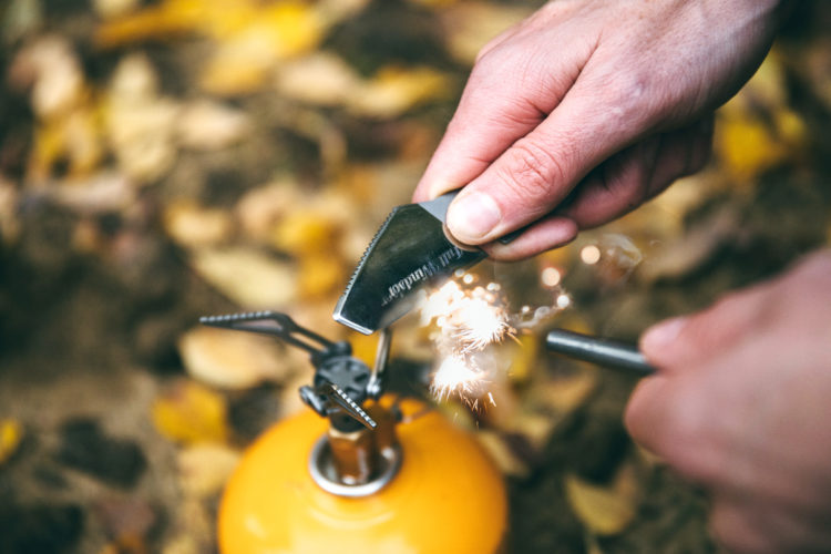 A close-up image showing a person's hand using a tool to create sparks on a camping stove, set against a background of autumn leaves. The stove is orange, and the tool has a serrated edge.