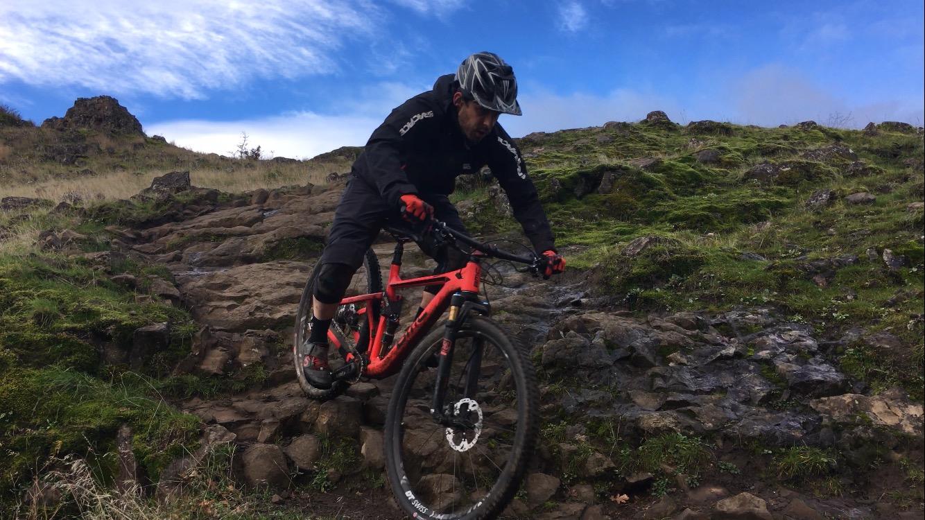 A mountain biker navigating a rocky trail on a bright day, focused and leaning forward on a vibrant red bicycle. The scenic background features lush green grass and a rugged landscape with patches of blue sky and clouds. Syncline mountain bike trail.