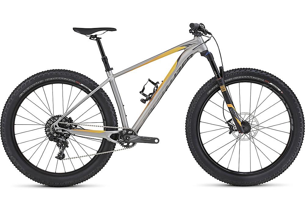 Specialized Fuse Comp 6Fattie: Alt text: A modern mountain bike with a silver frame and orange accents, featuring wide tires, a front suspension fork, and a single front chainring. The bike is positioned at an angle to showcase its design and components.