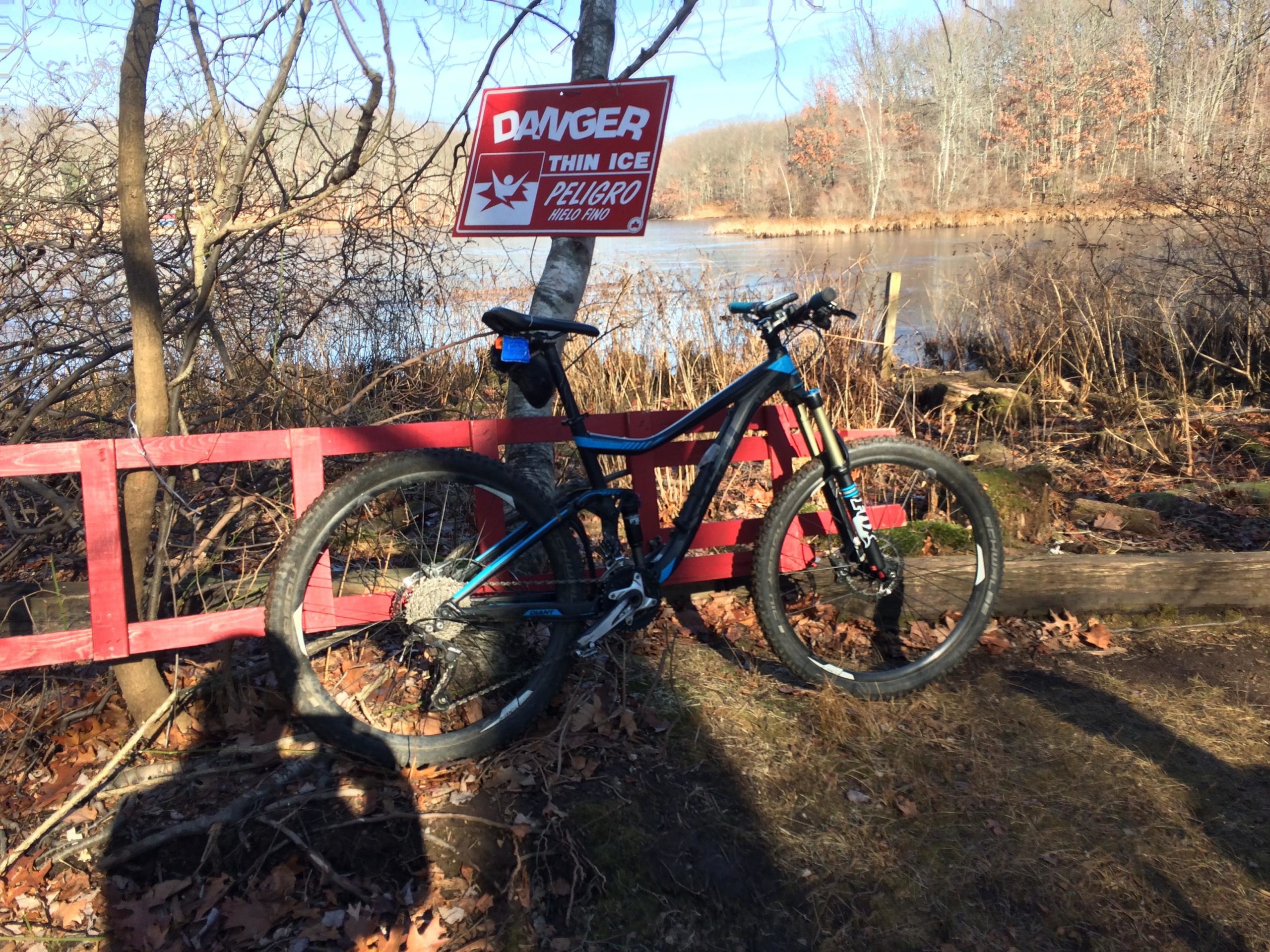A mountain bike rests against a red wooden fence near a body of water. A warning sign reading "Danger: Thin Ice" is visible in the background. The surrounding area features bare trees and fallen leaves, indicating a late autumn or early winter setting. Richmond Avenue and Forest Hill road mountain bike trail.