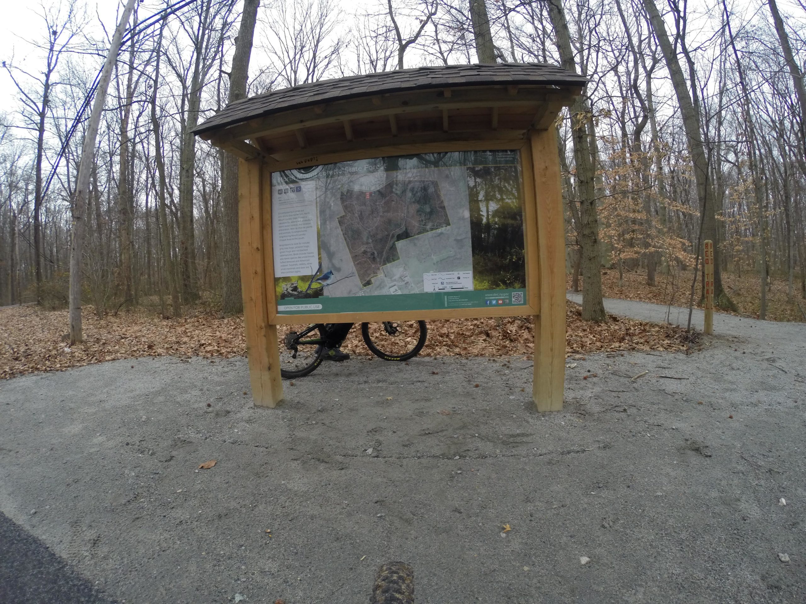 A wooden information kiosk with a trail map stands in a wooded area with bare trees and fallen leaves. A bicycle is positioned beside the kiosk, and a gravel path leads into the forest. The scene suggests a recreational area for hiking or biking. Long Pond mountain bike trail.