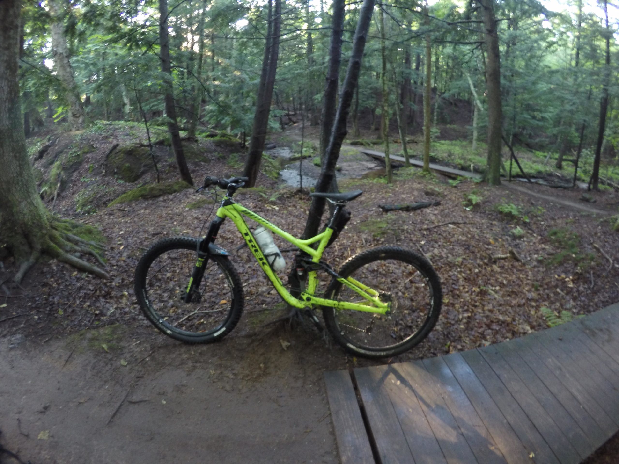 A bright yellow mountain bike stands on a dirt path in a forested area, surrounded by trees and lush greenery. The ground is covered in a mix of dirt and fallen leaves, and there is a wooden bridge visible in the background. Noquemanon Trails Network: South Marquette Trails mountain bike trail.