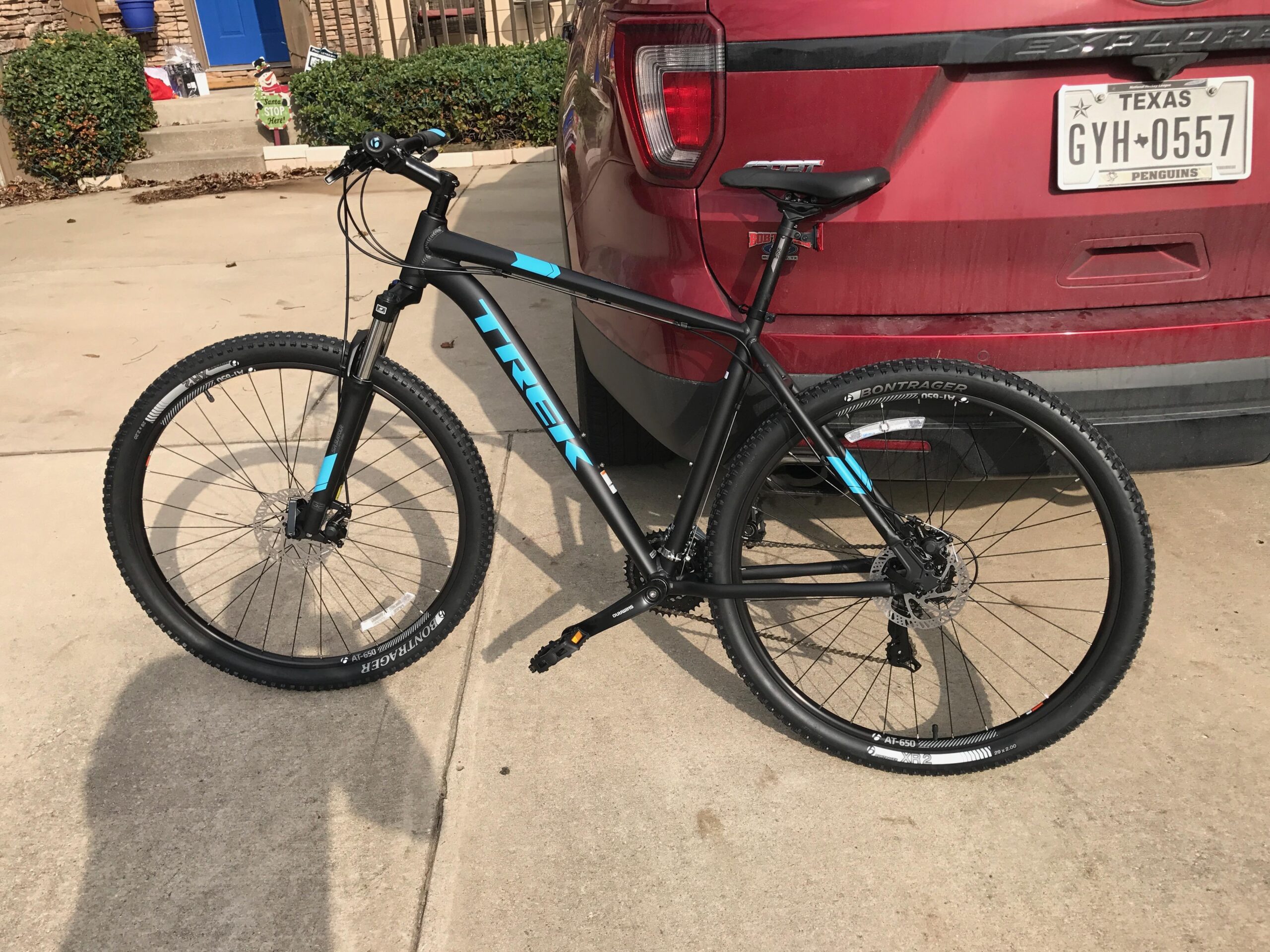 Trek Marlin 5: A black Trek mountain bike with blue accents leaning against a red SUV on a concrete driveway. The bike features a front suspension fork and rugged tires, with the background showing steps leading to a house.