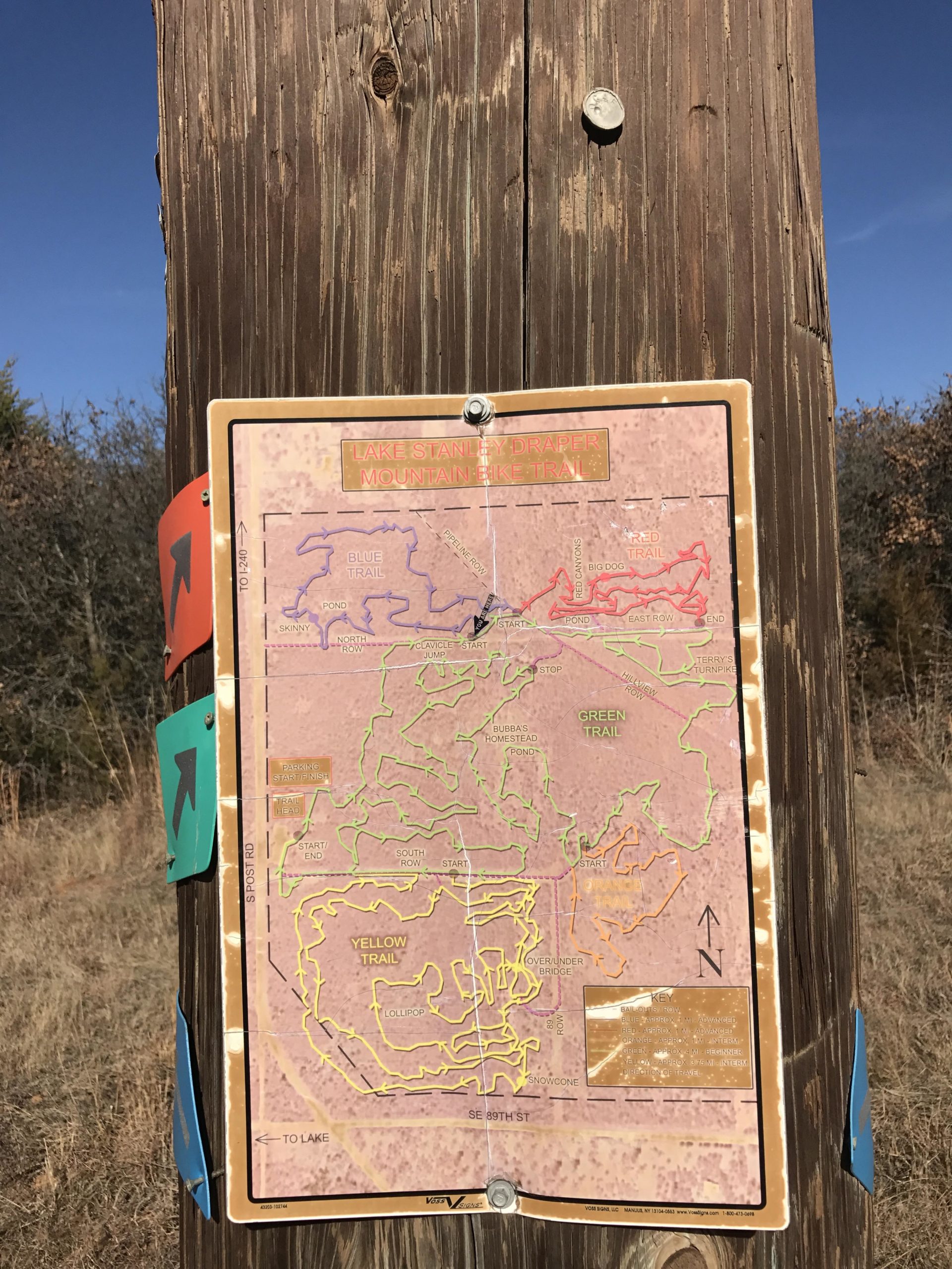 A wooden post displaying a trail map for Lake Stanley Draper Mountain Bike Trail. The map includes routes marked in different colors such as blue, red, green, and yellow, with labeled trails and landmarks. Surrounding the post is a natural landscape, and a clear blue sky is visible above. Lake Stanley Draper mountain bike trail.