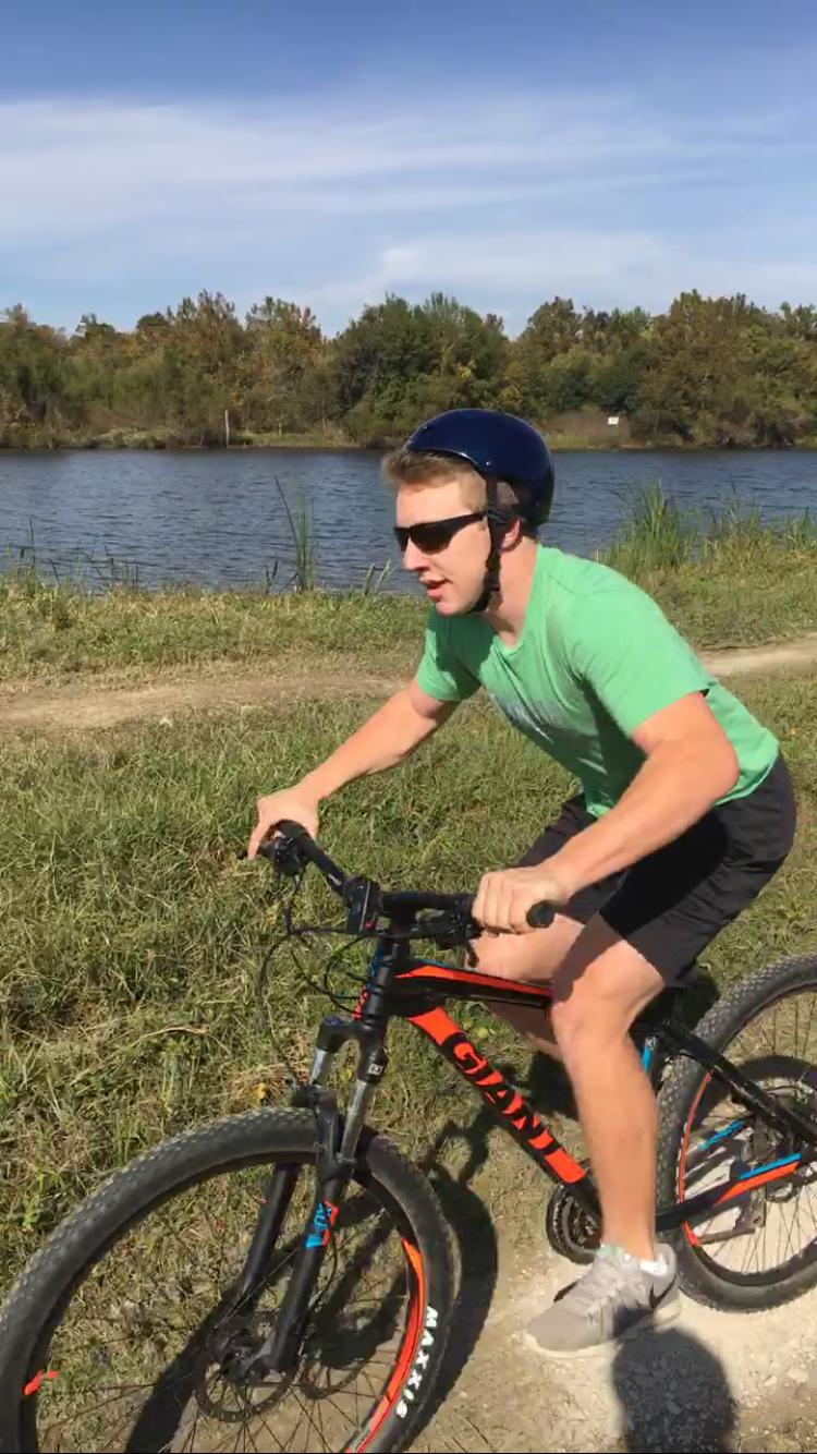 2017 Giant Talon 3: A young male cyclist wearing a blue helmet and sunglasses rides a black and orange mountain bike along a dirt path next to a calm lake, surrounded by greenery under a clear blue sky.