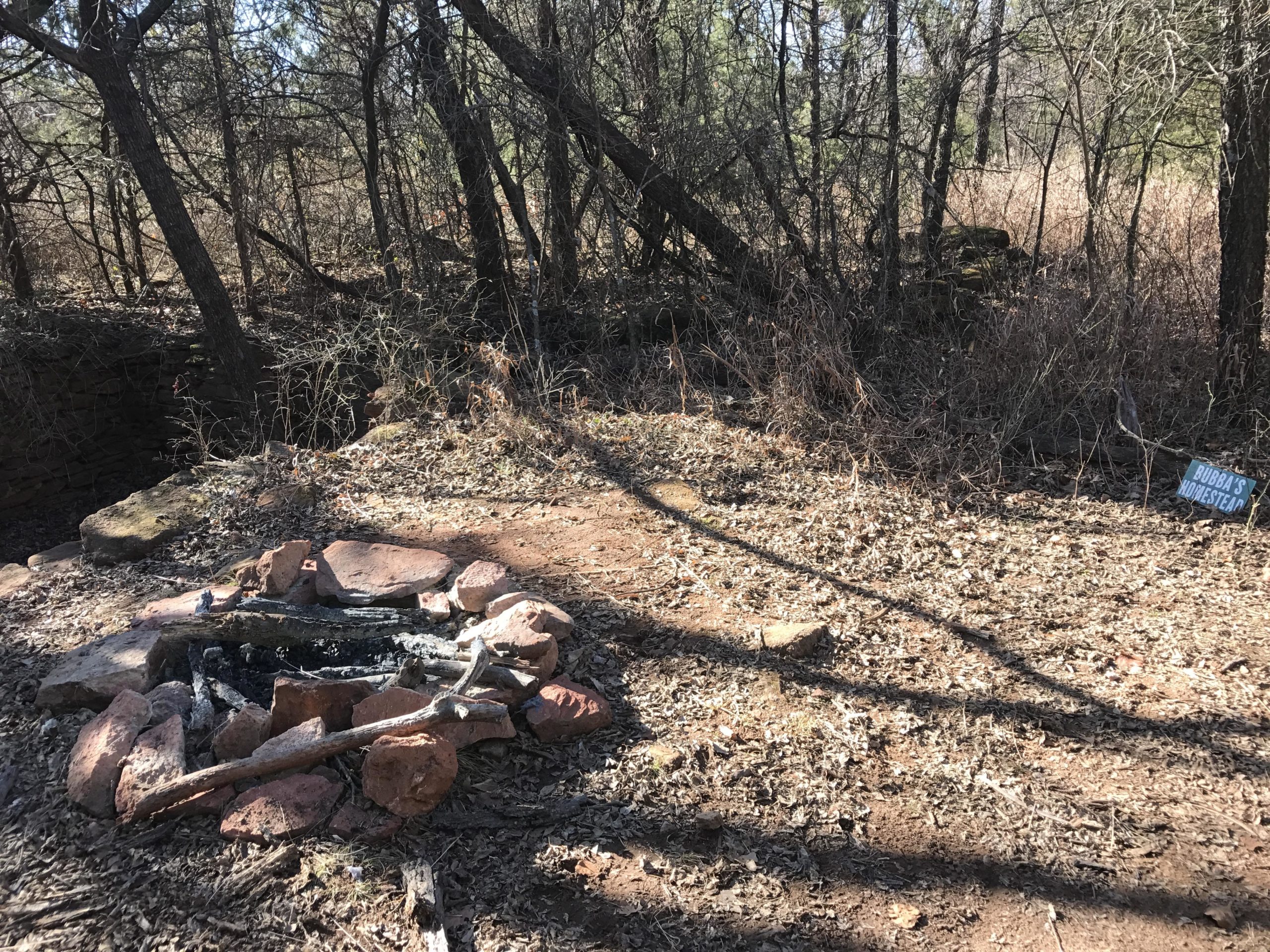 A campsite area featuring a stone fire pit surrounded by scattered leaves and twigs. In the background, there are dense trees and underbrush. A sign reading "Bubba's Homestead" is visible to the right of the fire pit. Lake Stanley Draper mountain bike trail.