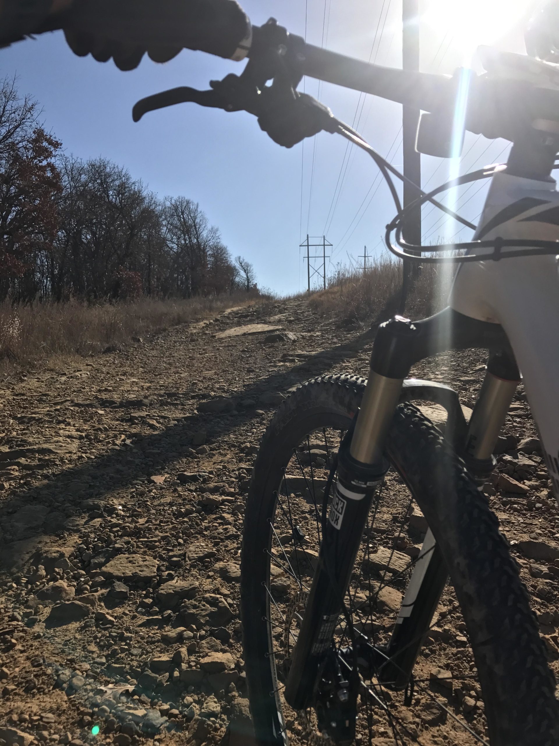 Close-up view of a mountain bike resting on a rocky trail, with the handlebars and front wheel in focus. The background features a clear blue sky and a sunburst effect, along with sparse tree lines and utility poles along the horizon, indicating an outdoor biking adventure. Turkey Mountain mountain bike trail.
