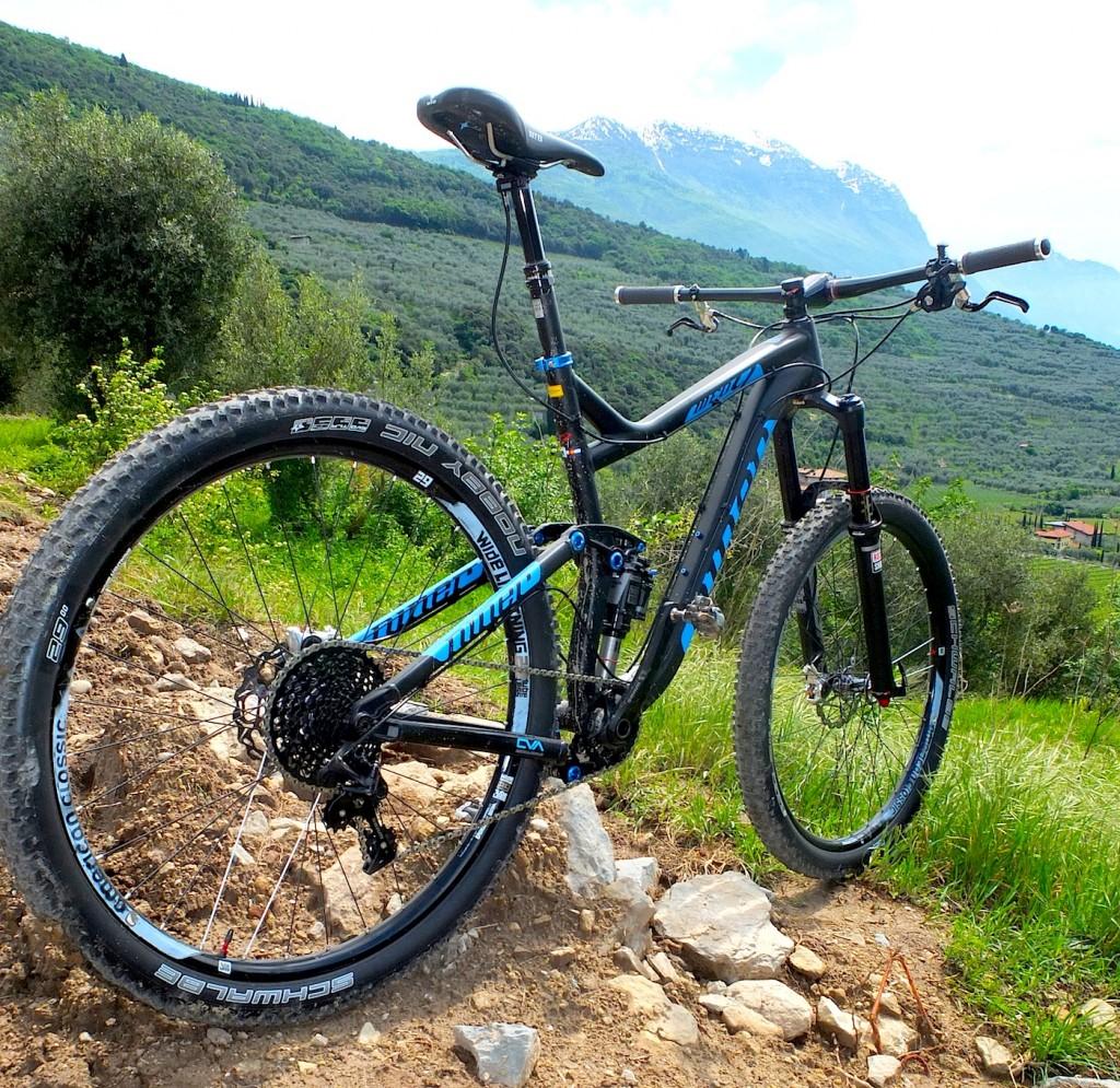A modern mountain bike positioned on a gravel path, surrounded by lush green hills and distant snow-capped mountains under a bright sky. The bike features a sleek black frame with blue accents, thick tires designed for rugged terrain, and a well-defined gear system visible on the rear. Nocatee mountain bike trail.