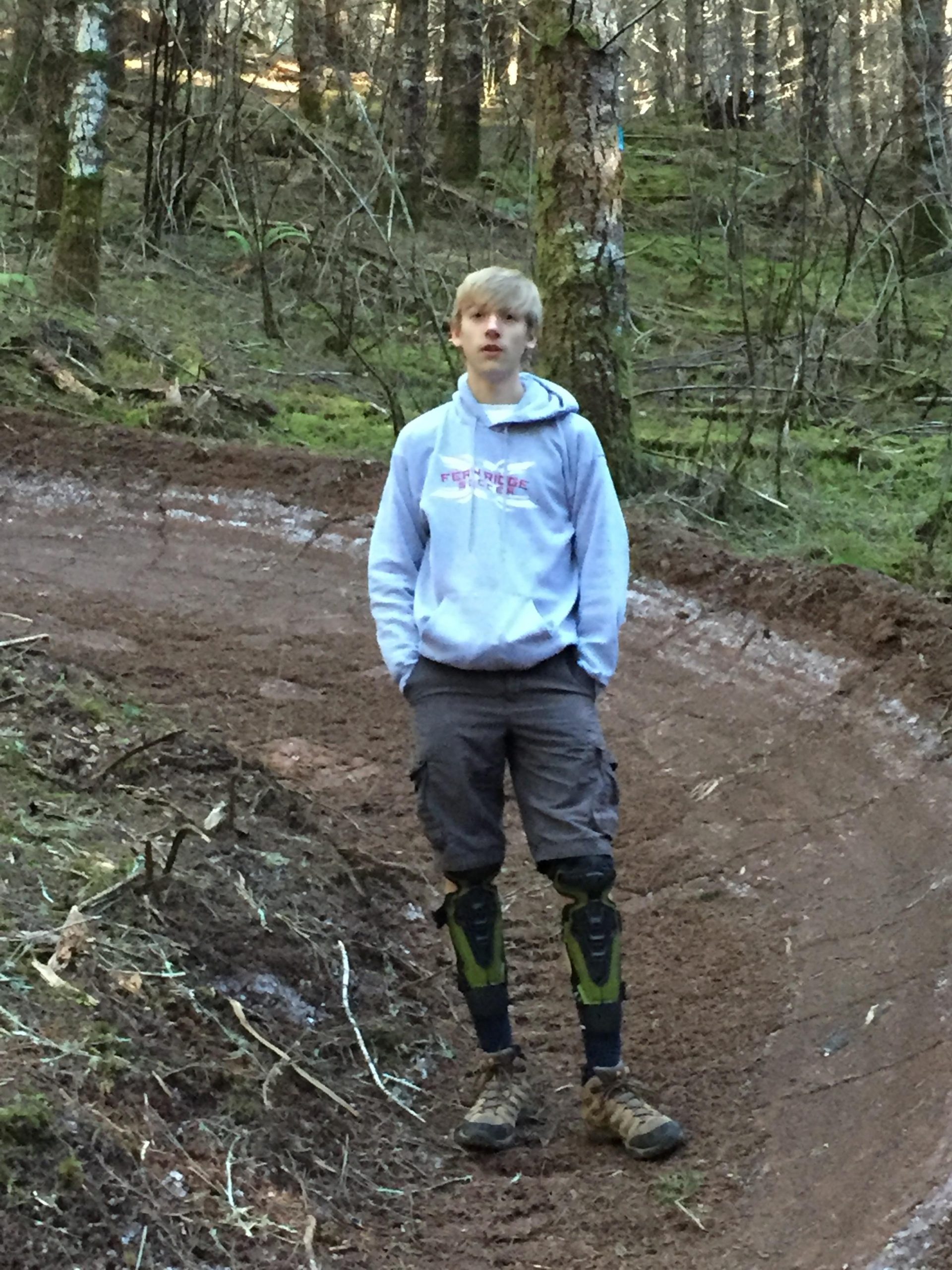A young person standing on a dirt path in a forested area, wearing a light gray hoodie, cargo shorts, and protective knee pads. The background features trees and green foliage, with sunlight filtering through the branches. The individual appears relaxed and is looking slightly off-camera. Whypass mountain bike trail.