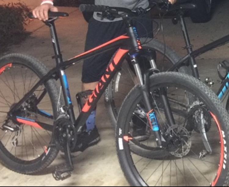 2017 Giant Talon 3: Two mountain bikes are parked side by side in a garage. One bike features a black frame with orange and blue accents, while the other bike has a predominantly black design. A person's legs wearing dark shorts and cycling shoes are visible beside the bikes. The background includes garage details, such as a vehicle and a well-lit space.