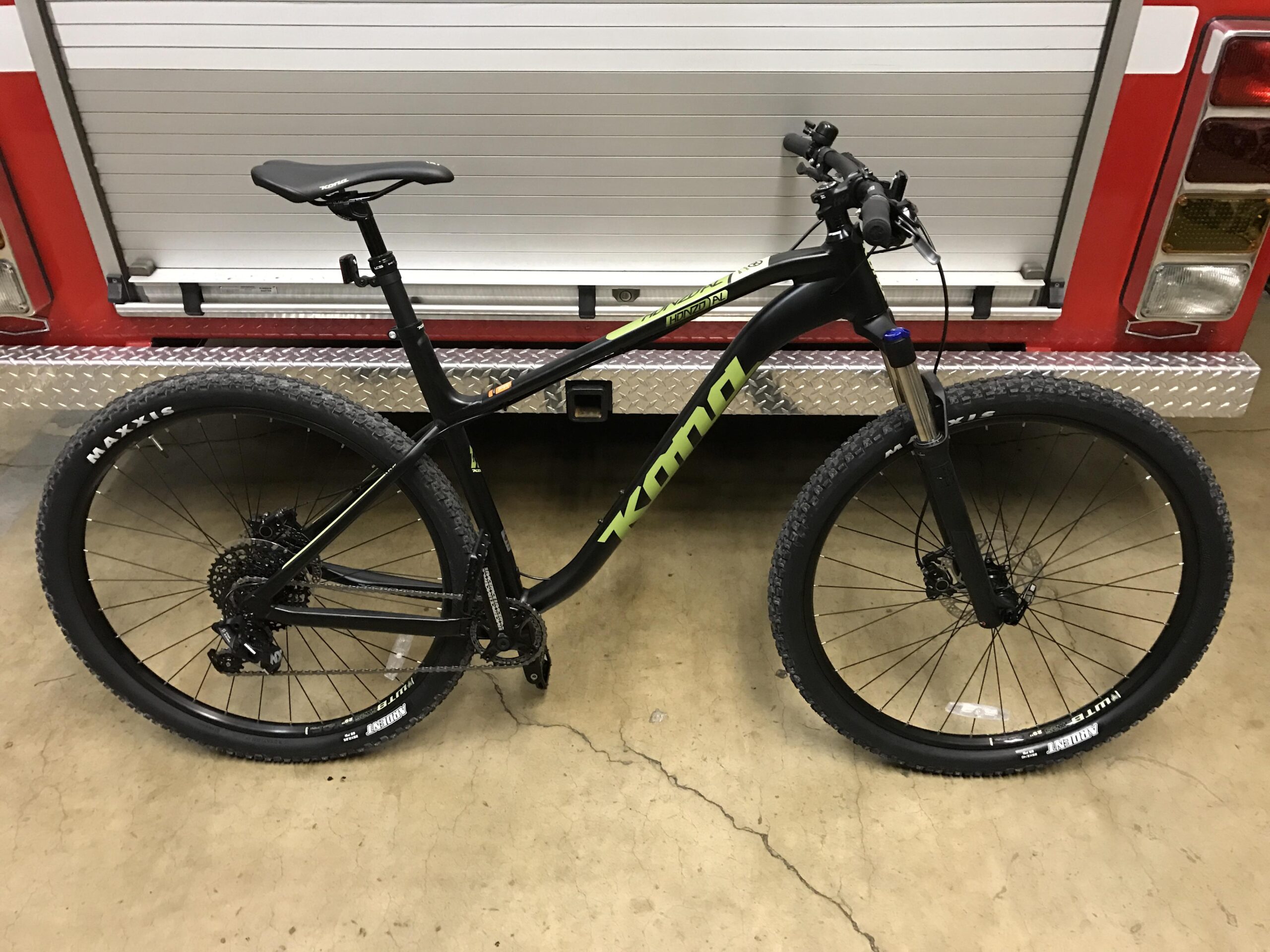 Kona Honzo: A black mountain bike with green accents parked in front of a fire truck. The bike features wide tires labeled "Maxxis," a front suspension fork, and a sleek design suitable for off-road riding. The background includes a red and silver fire truck, showcasing a diamond plate surface.