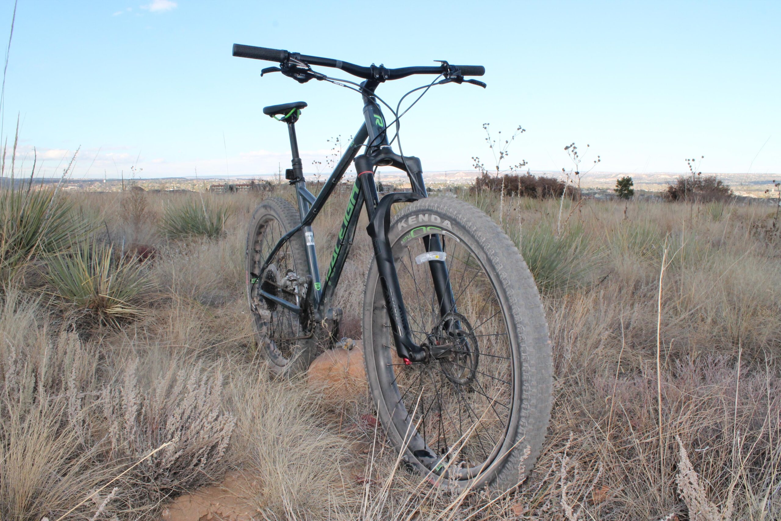 Raleigh Tokul 4130: A black mountain bike is positioned in a grassy landscape, surrounded by sparse vegetation. The bike features wide tires and a modern frame design, with the handlebars angled towards the viewer. In the background, a distant view of rolling hills and an open sky can be seen.