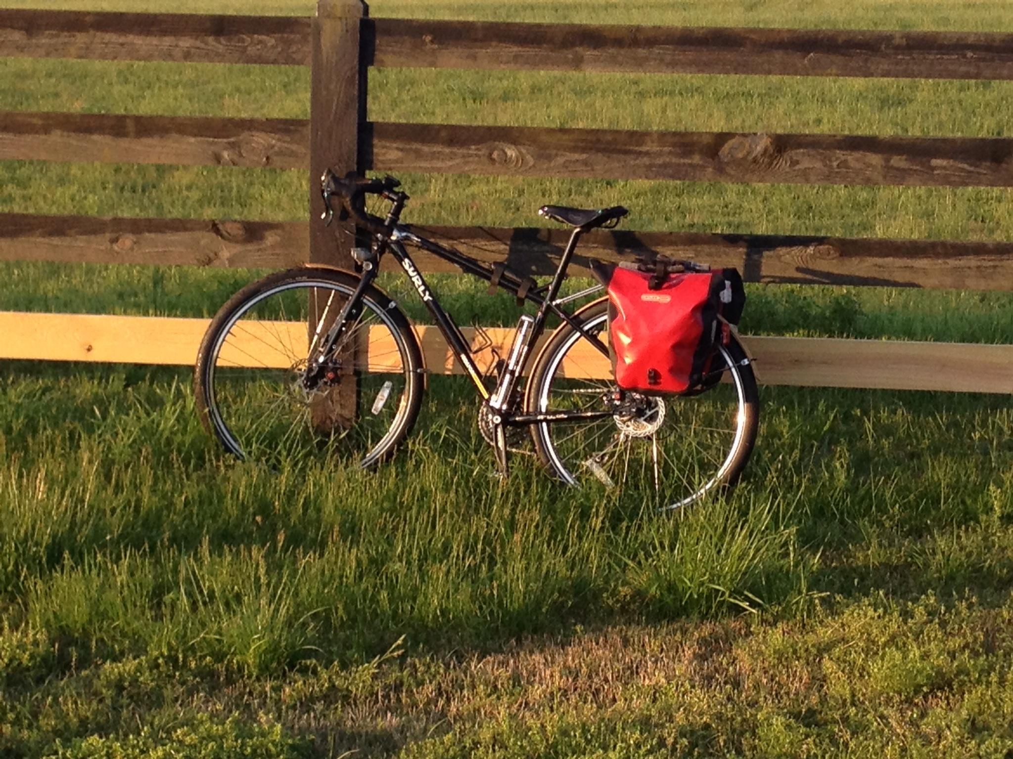 Surly Ogre: A black bike with a red saddlebag parked next to a wooden fence on a grassy field. The scene is illuminated by warm sunlight, creating a peaceful outdoor atmosphere.