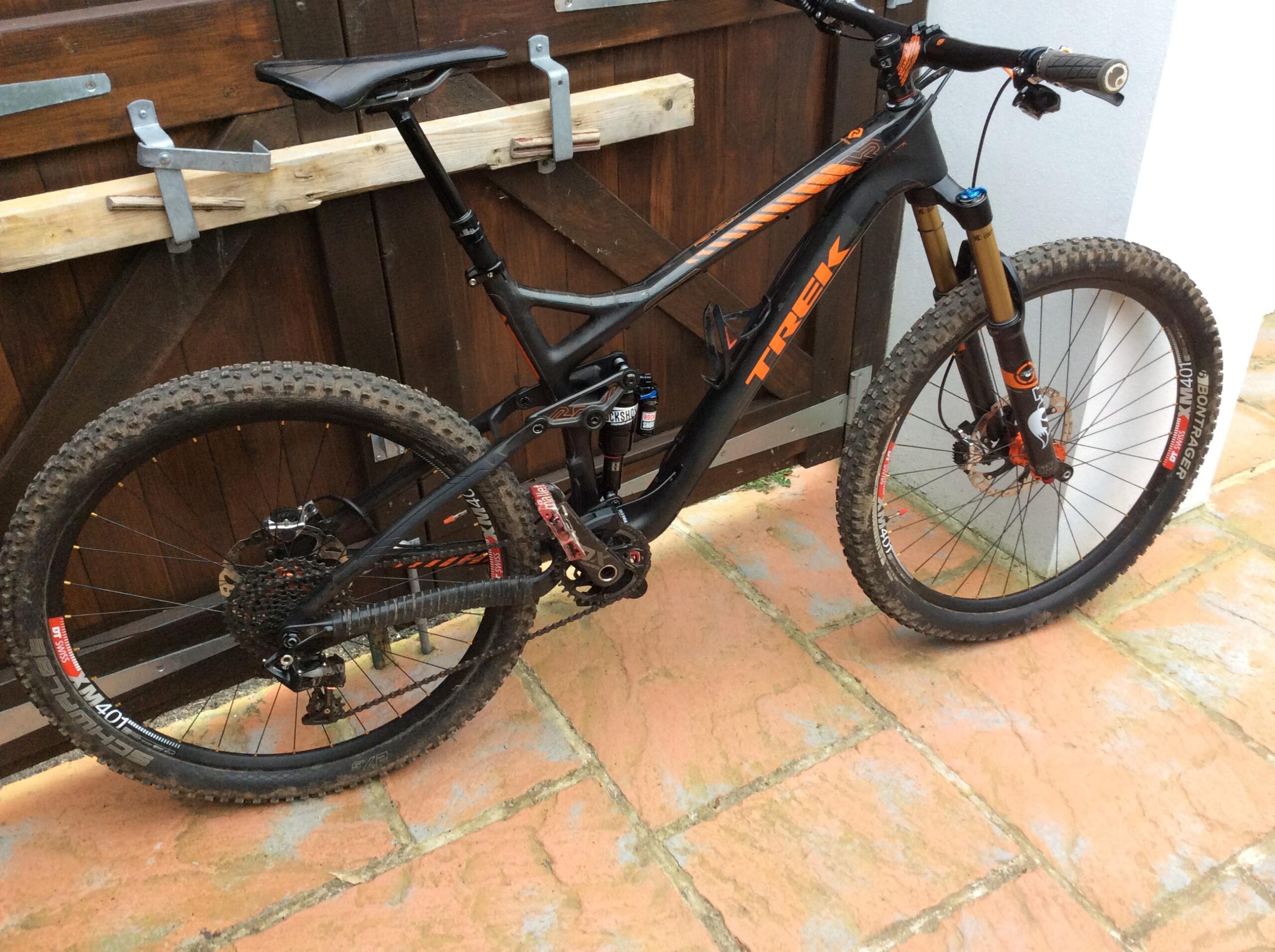 Trek Remedy: A black and orange mountain bike leaning against a wooden gate, featuring a lightweight frame, knobby tires, and visible suspension components. The bike rests on a tiled surface, showcasing its tires with some dirt, indicating recent use.
