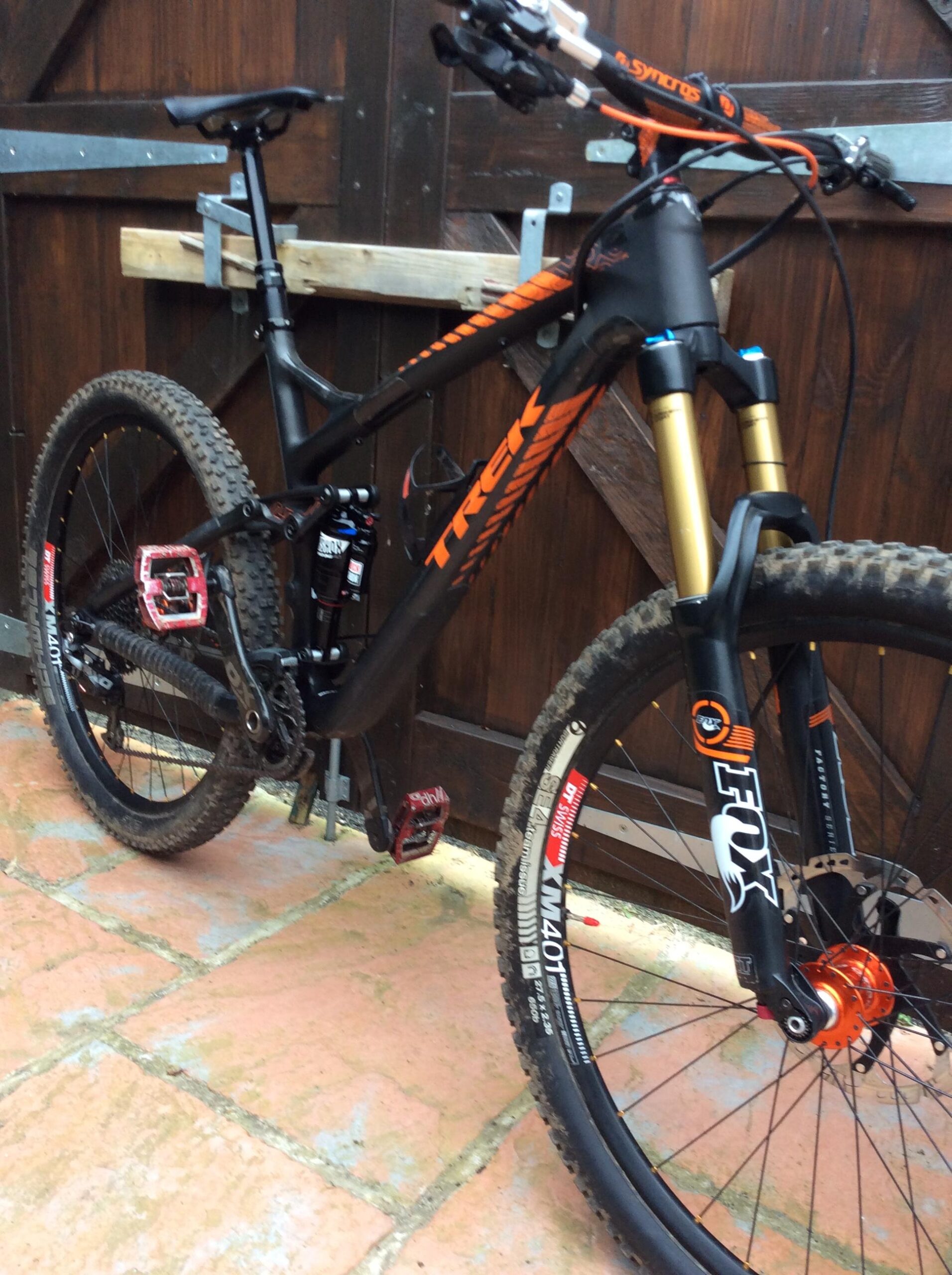 Trek Remedy: A close-up view of a mountain bike featuring a sleek black and orange frame, equipped with front suspension forks, disc brakes, and rugged tires. The bike is positioned against a wooden backdrop, showcasing its dirt-streaked tires indicating recent use.