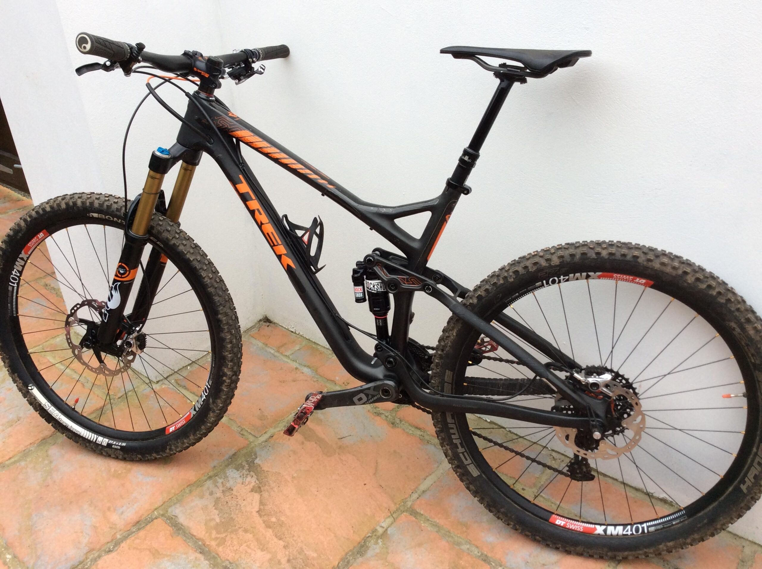 Trek Remedy: A black and orange Trek mountain bike leaning against a white wall, with prominent features such as a front suspension, wide tires with knobby tread, and a dropper seat post. The bike rests on a tiled surface, showcasing its detailed components like the gear system and disc brakes.