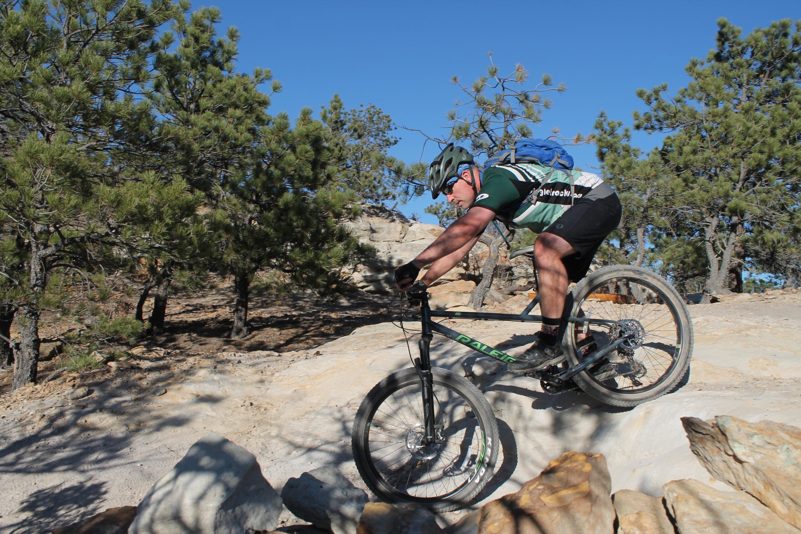 Raleigh Tokul 4130: A mountain biker navigating rocky terrain, leaning forward on their bike, with trees in the background and a clear blue sky overhead. The cyclist is wearing a helmet and a green and black jersey, with a backpack secured on their back.