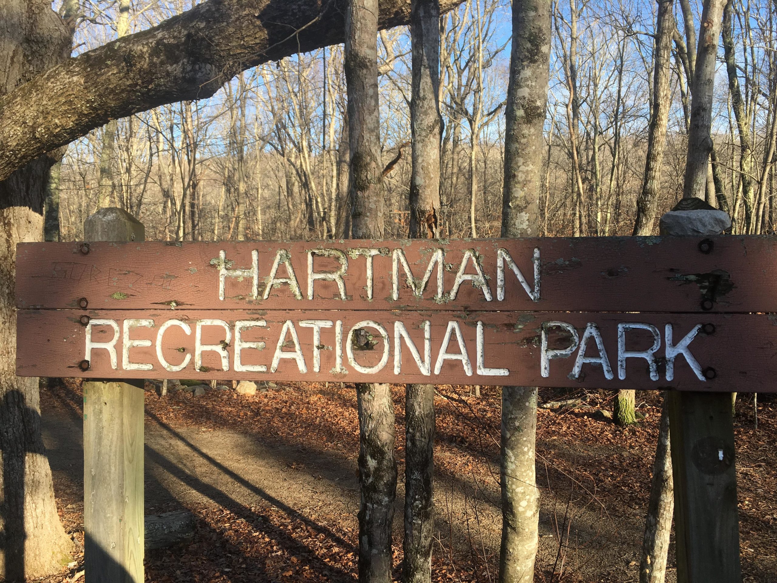 A close-up view of a wooden sign that reads "Hartman Recreational Park," mounted on a post, surrounded by bare trees and fallen leaves in a natural setting. Hartman Park mountain bike trail.