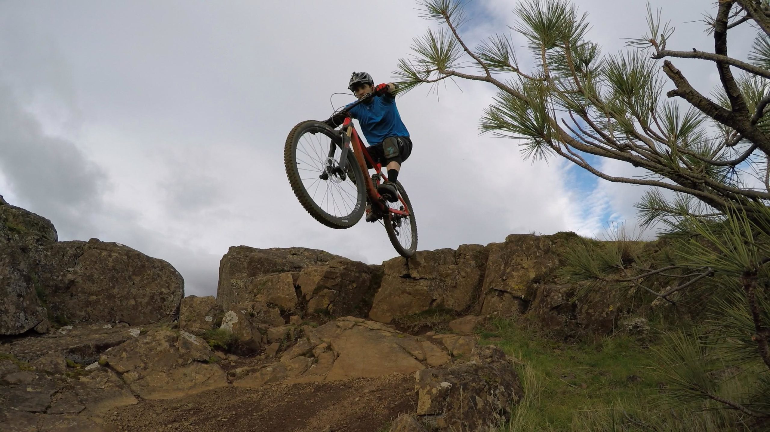 A mountain biker performs a jump over rocky terrain, with one wheel lifted off the ground and a tree in the background against a cloudy sky. Syncline mountain bike trail.