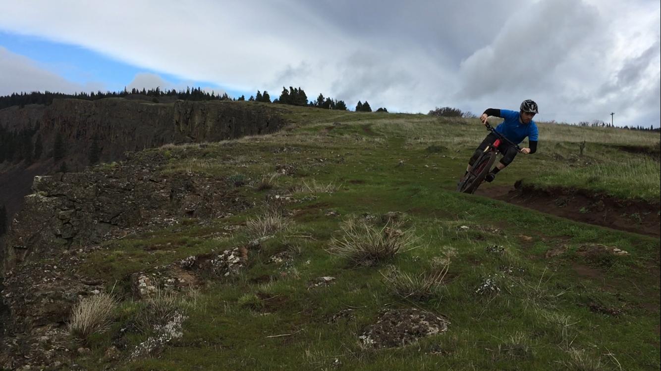 A mountain biker wearing a blue shirt and helmet leans into a turn on a grassy hillside, with a rocky cliff and trees in the background under a cloudy sky. Syncline mountain bike trail.