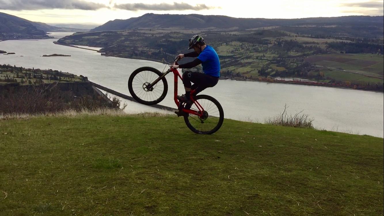 A mountain biker performs a wheelie on a grassy hillside overlooking a river, with scenic hills and cloudy skies in the background. Syncline mountain bike trail.