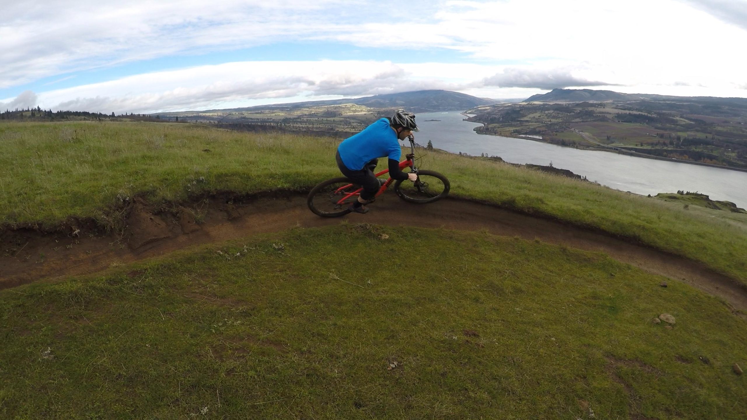 A mountain biker rides along a winding dirt path on a grassy hillside, with a scenic view of a river and rolling hills in the background under a partly cloudy sky. Syncline mountain bike trail.