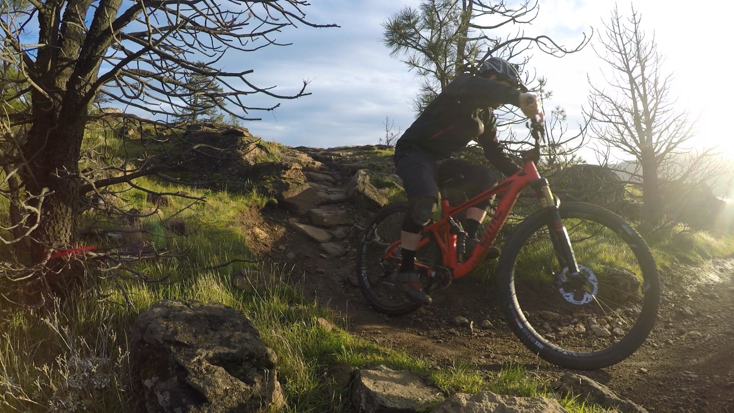 A mountain biker navigating a rocky trail surrounded by grass and sparse trees under a partly cloudy sky. The biker is wearing a helmet and riding a bright red mountain bike. Sunlight is filtering through the trees, creating a dynamic outdoor scene. Syncline mountain bike trail.