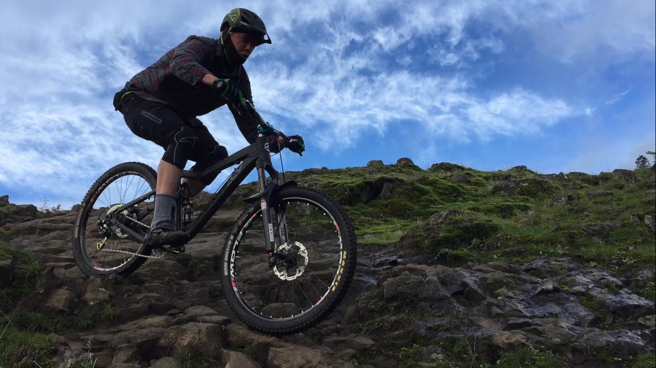 A person riding a mountain bike down a rocky terrain with grass and moss, under a bright blue sky with scattered clouds. The rider is wearing a helmet and protective gear, demonstrating a dynamic position as they navigate the challenging landscape. Syncline mountain bike trail.