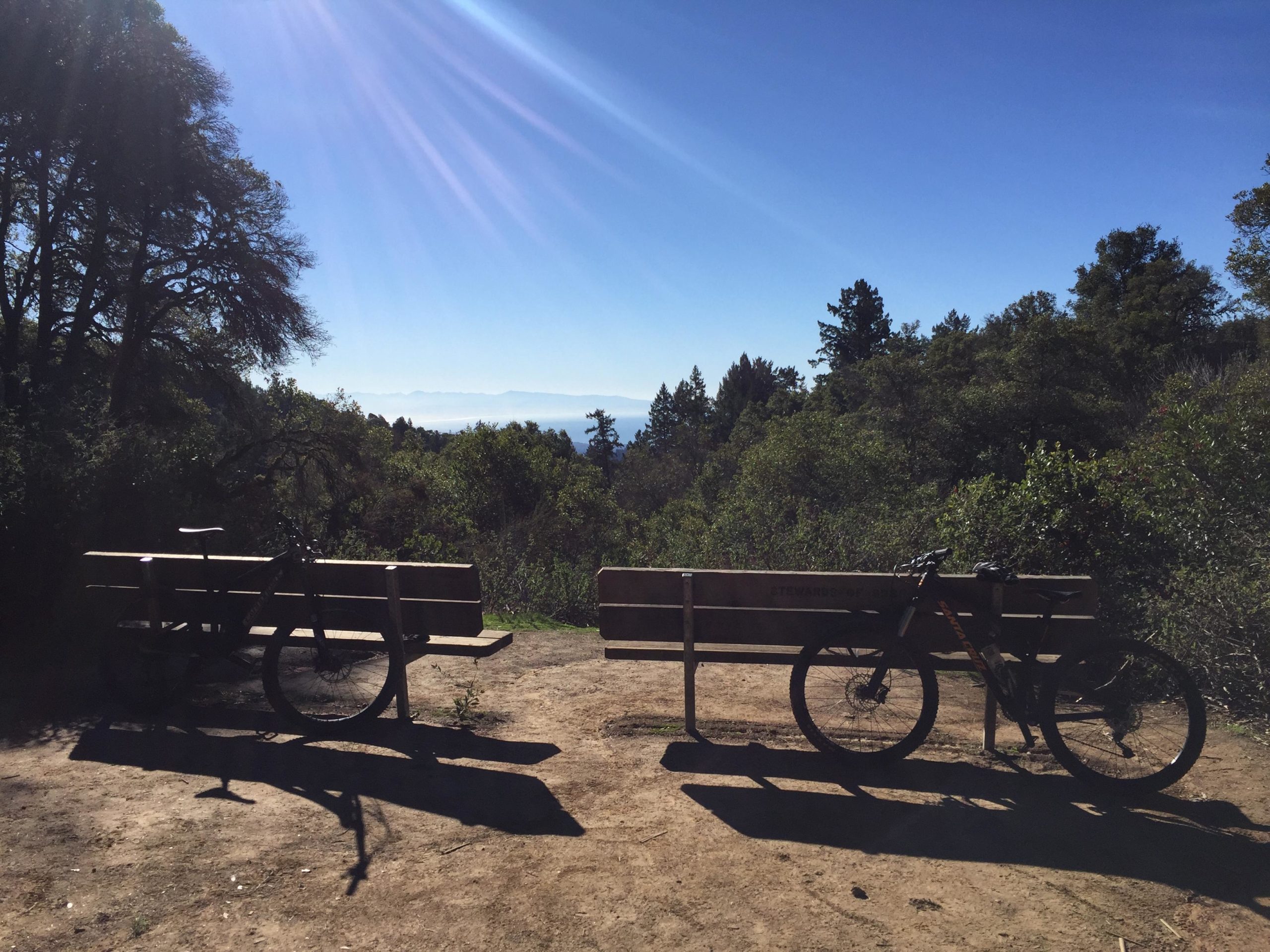 Two mountain bikes parked beside wooden benches in a sunlit outdoor setting, surrounded by trees and greenery. Clear blue sky with rays of sunlight shining down, creating a serene atmosphere. Forest Of Nisene Marks and Soquel Demonstration Forest mountain bike trail.
