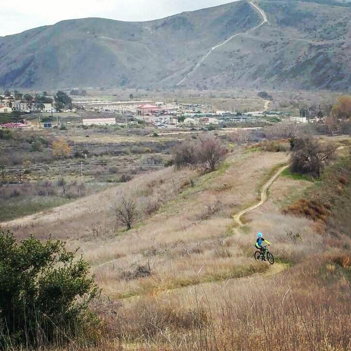 A mountain biker rides along a dirt trail in a grassy landscape, surrounded by rolling hills and distant mountains. Sparse trees and shrubs are visible, with a small town nestled in the valley below. The sky is overcast, creating a serene outdoor scene. San Clemente Singletracks mountain bike trail.