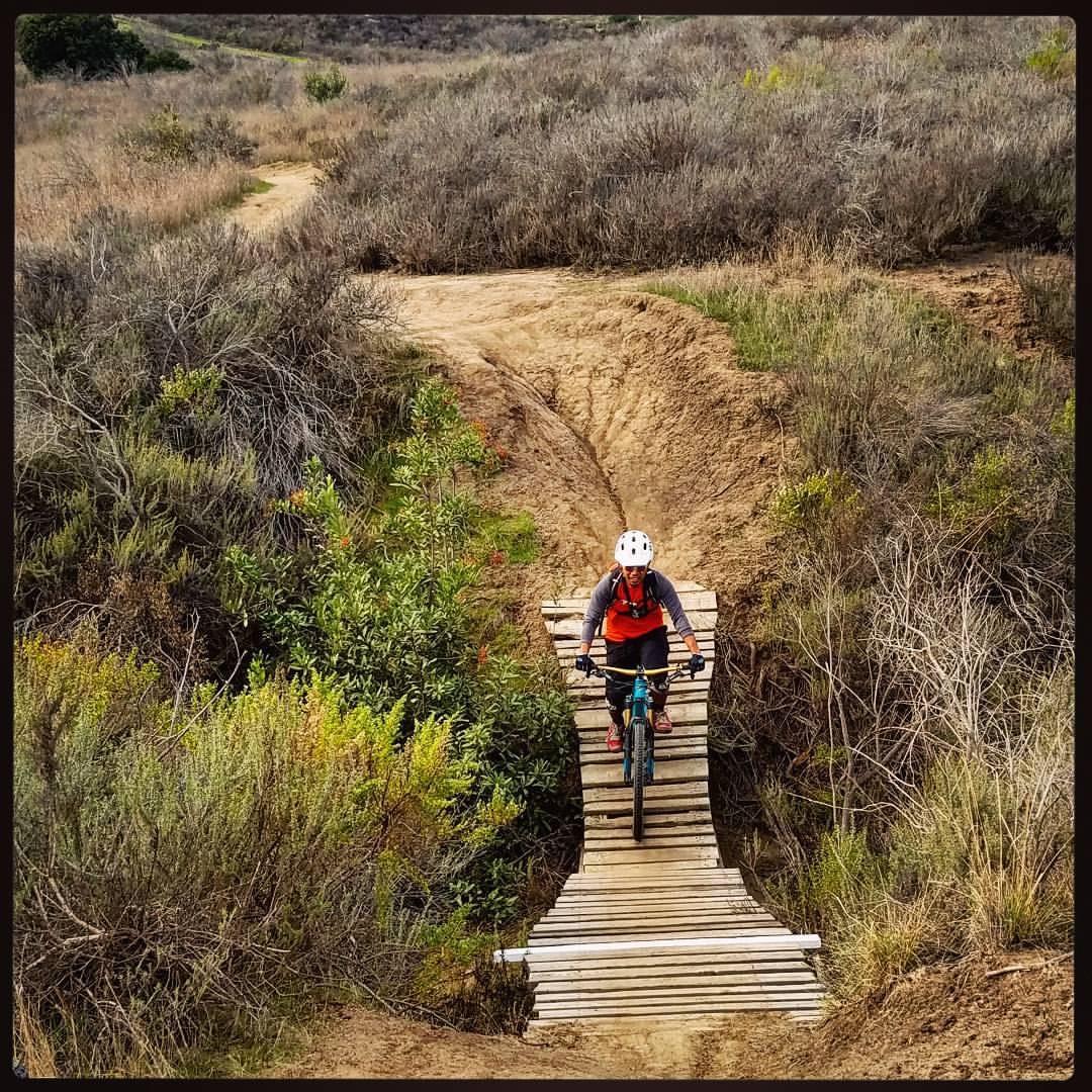 A person riding a mountain bike on a wooden bridge over rough terrain, surrounded by shrubs and dry grasses in a natural landscape. San Clemente Singletracks mountain bike trail.