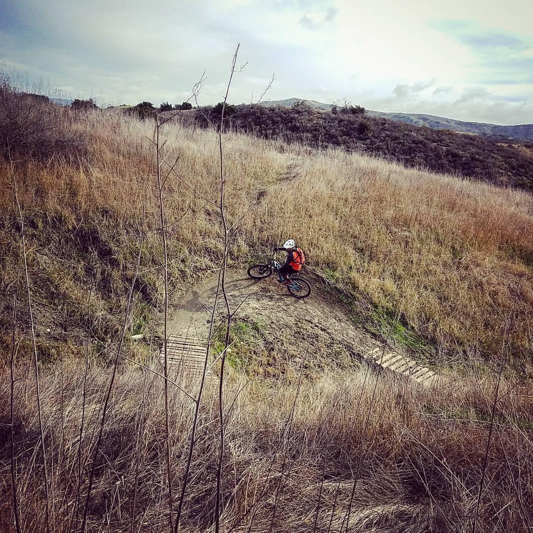 A cyclist in a red jacket rides a mountain bike along a winding dirt trail surrounded by tall dry grass and hills under a cloudy sky. San Clemente Singletracks mountain bike trail.