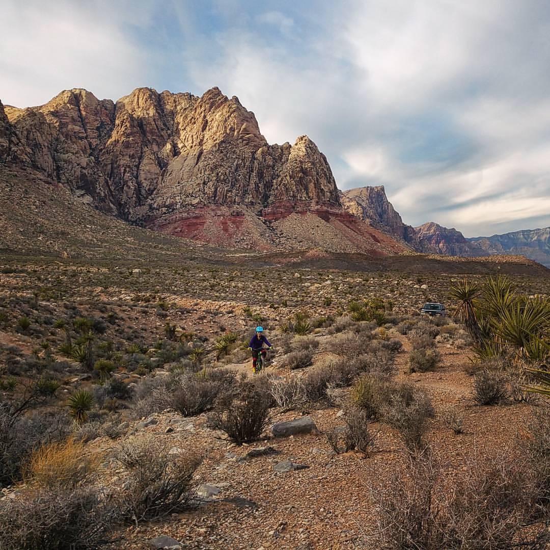 A person cycling on a dirt path in a desert landscape, with large rock formations in the background and a vehicle parked nearby. Sagebrush and other desert vegetation are scattered across the foreground, under a partly cloudy sky. Cottonwood Valley North mountain bike trail.