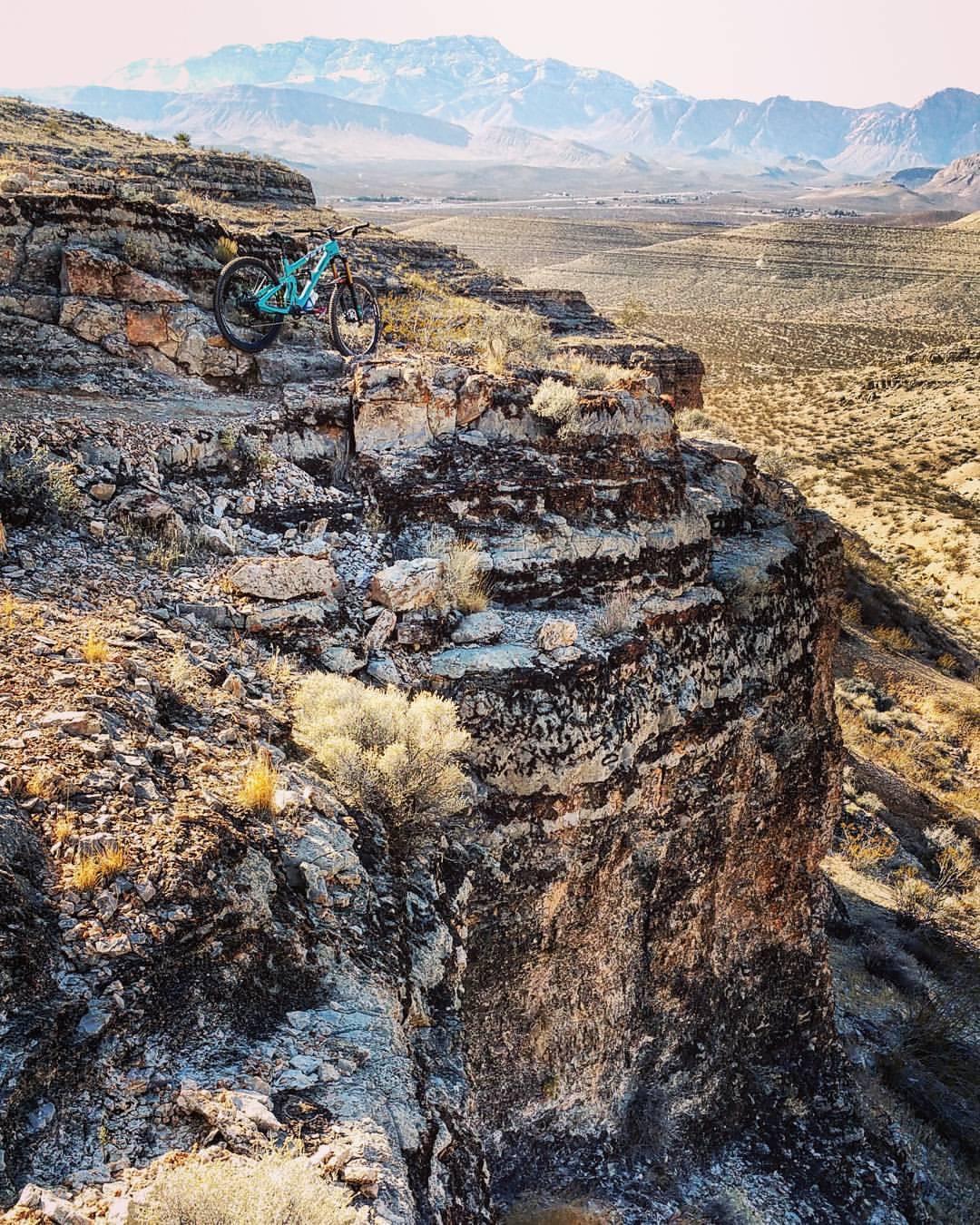 A mountain bike rests on the edge of a rocky cliff, overlooking an expansive landscape with mountains in the background and a desert terrain below. The scene captures the beauty of nature and the thrill of outdoor adventure. Ikes peak mountain bike trail.