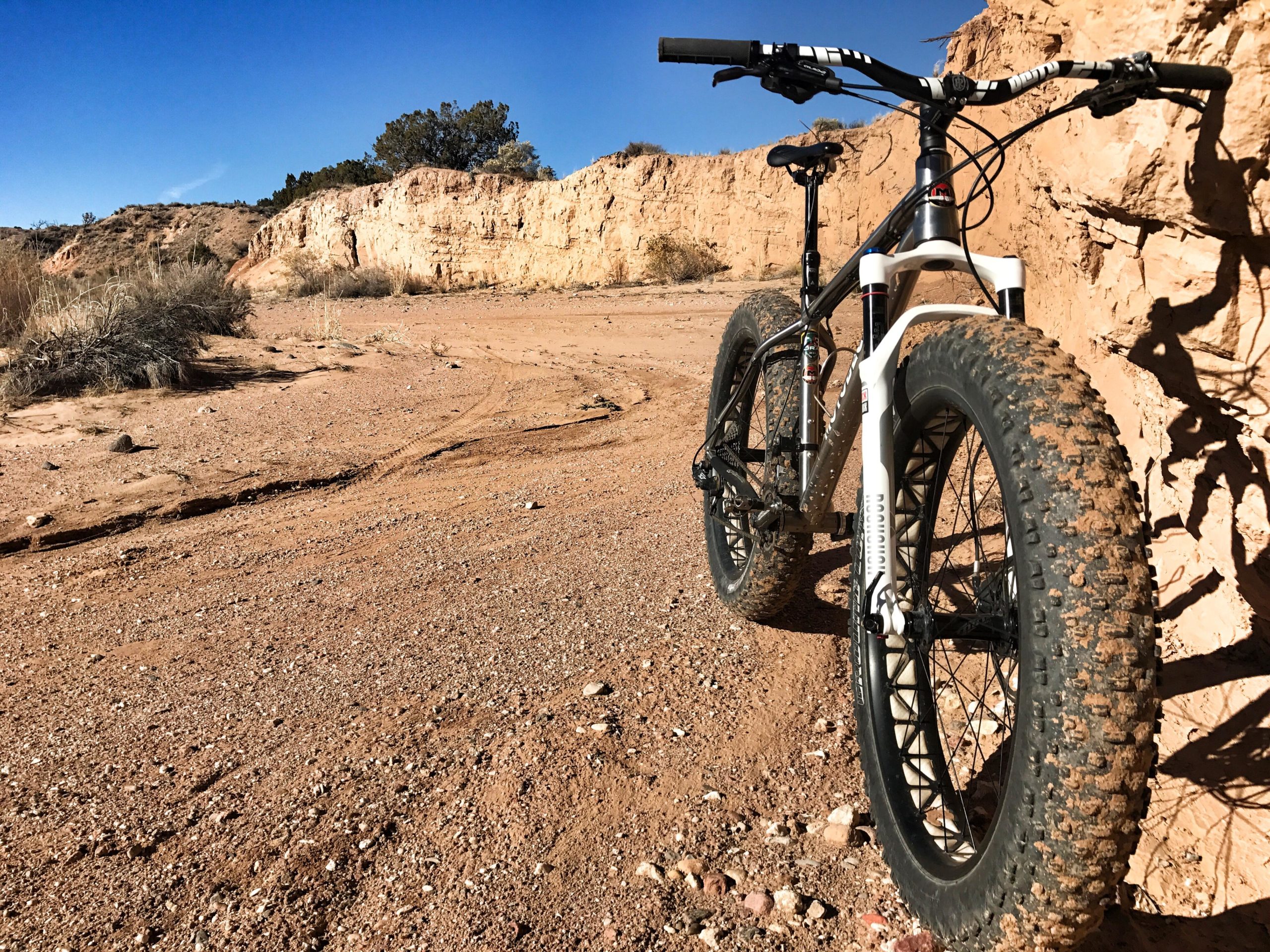 A close-up view of a fat bike resting on a sandy dirt trail, with rocky terrain and desert vegetation in the background. The sun casts a clear blue sky overhead, highlighting the bike's large tires and rugged frame. Mariposa Fat Bike Trails mountain bike trail.